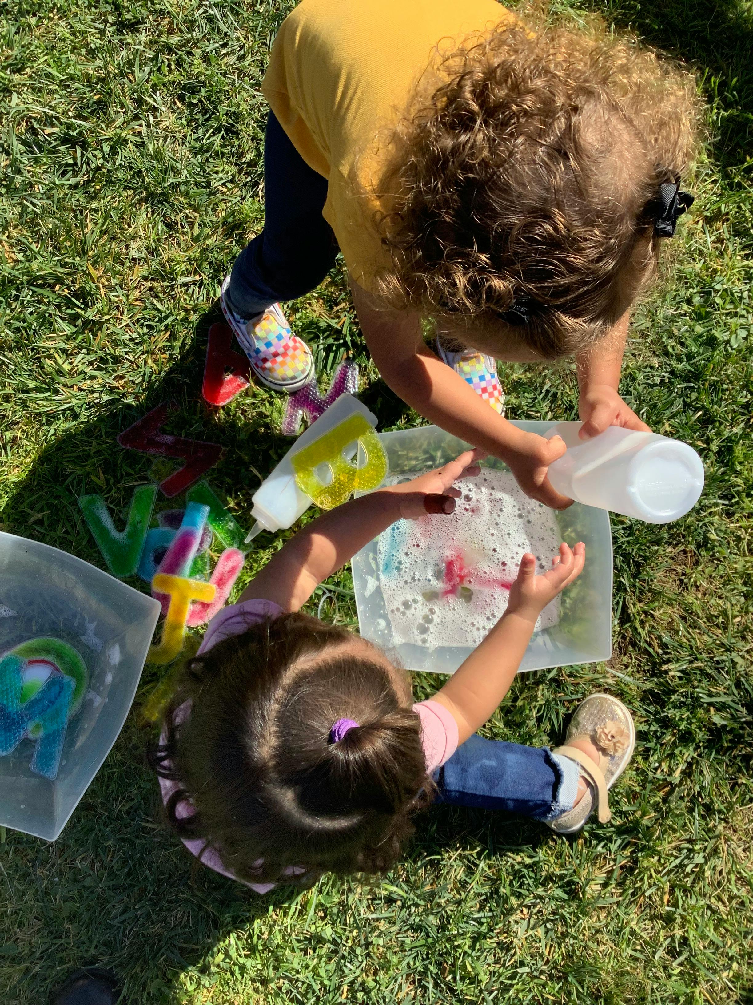 Two children playing with water and bubbles in plastic containers on grass