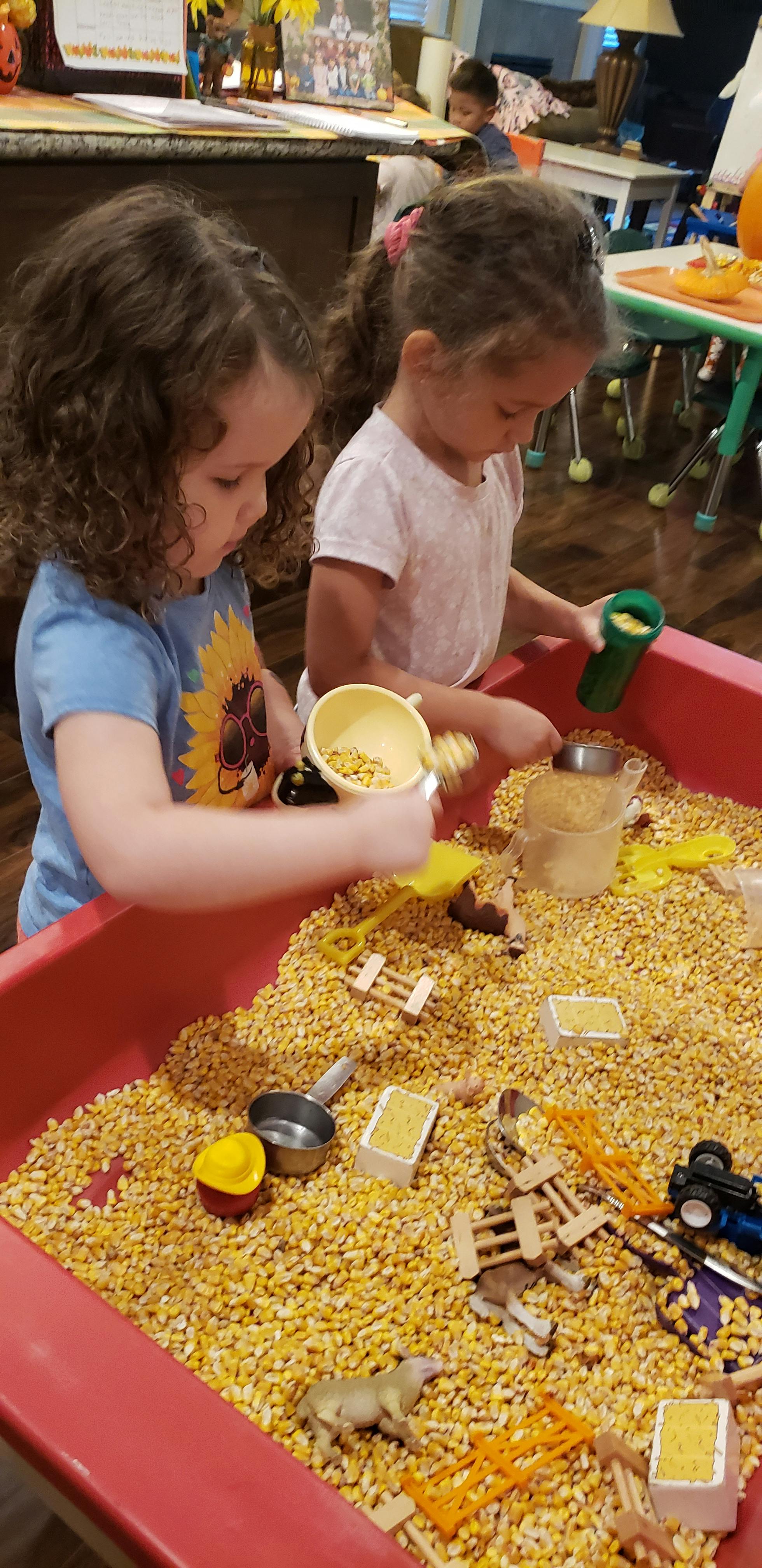 Two children playing with corn kernels and small toys in a red bin