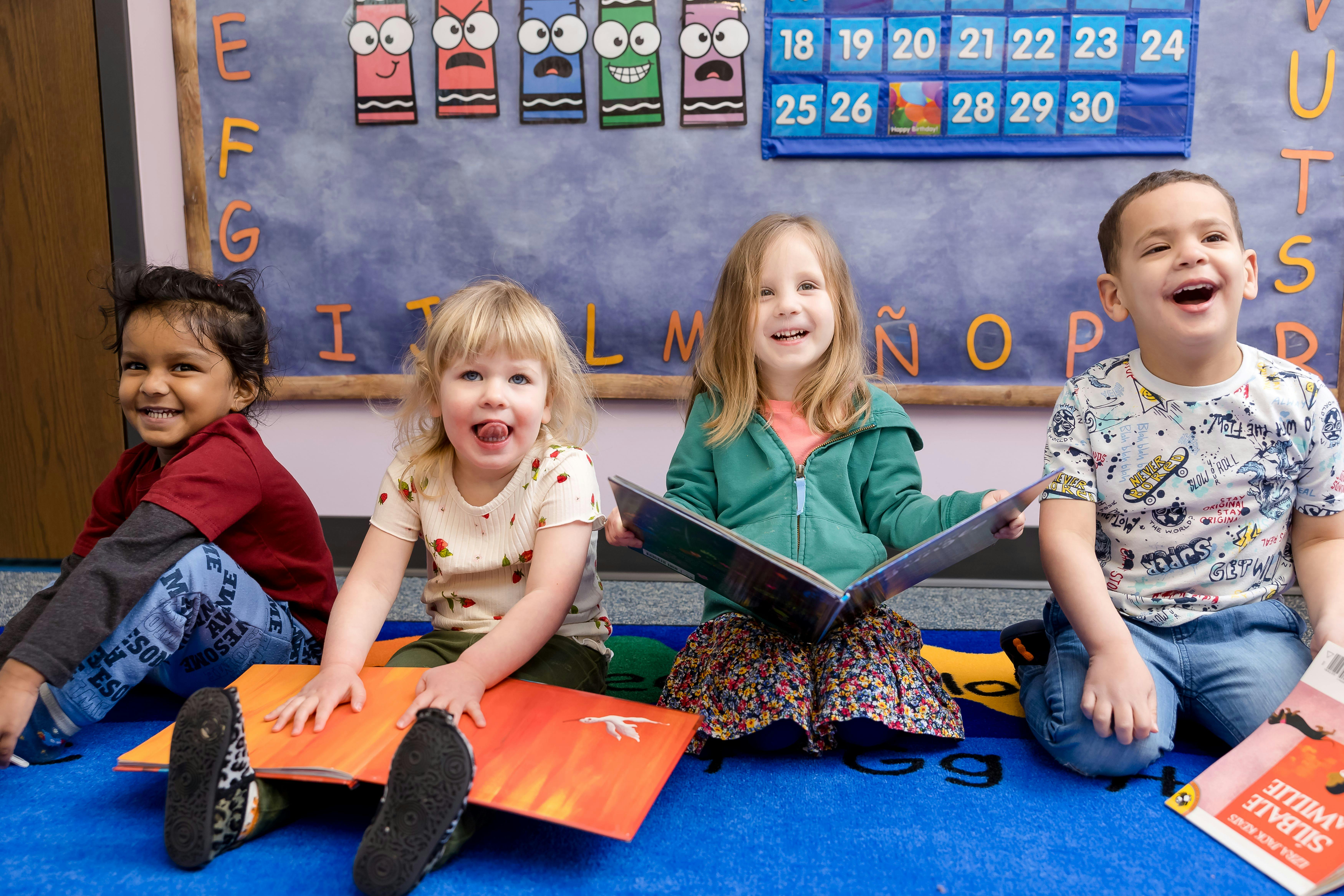 Four children sitting on the floor in a classroom, two holding books