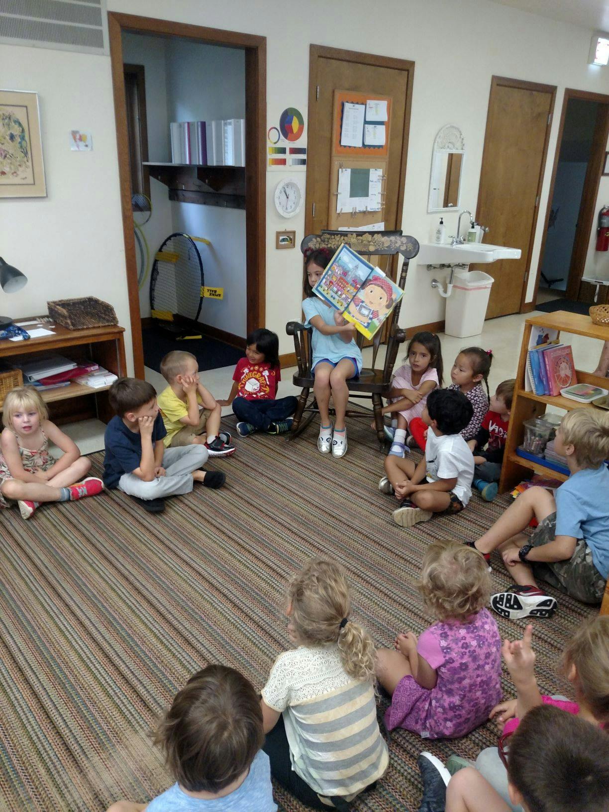 Child sitting in a chair reading a book to a group of children on the floor