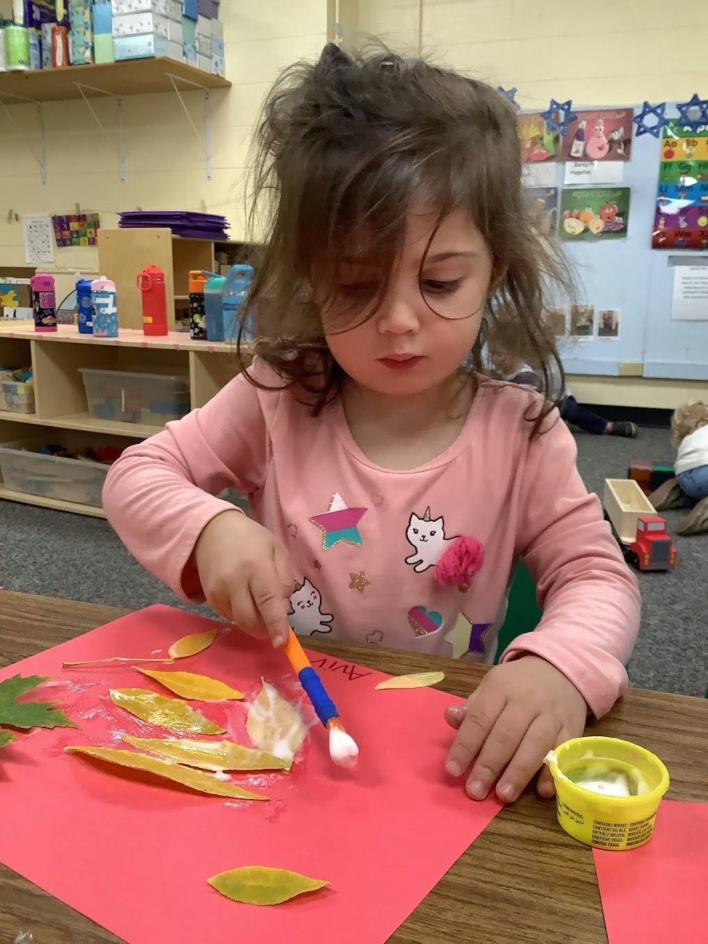 Child applying glue to leaves on red paper in a classroom