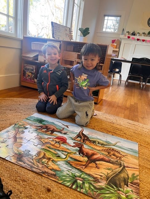 Two children kneeling behind a dinosaur puzzle in a classroom