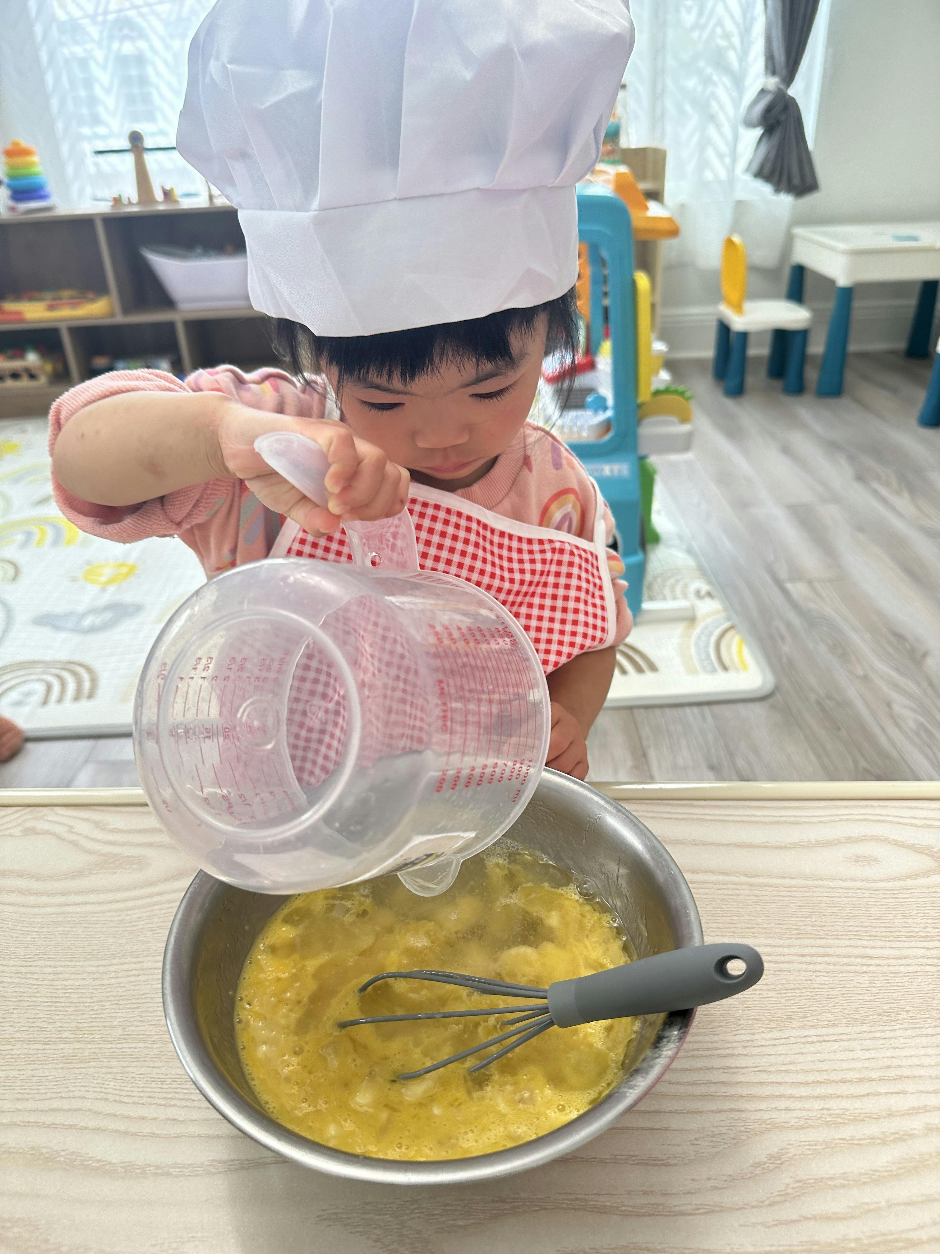 Child wearing a chef hat and apron pouring liquid into a metal mixing bowl