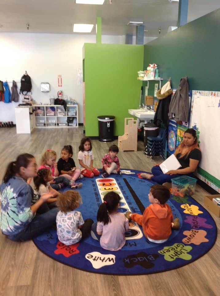 Two adults and multiple children sitting in a circle on a rug in a classroom
