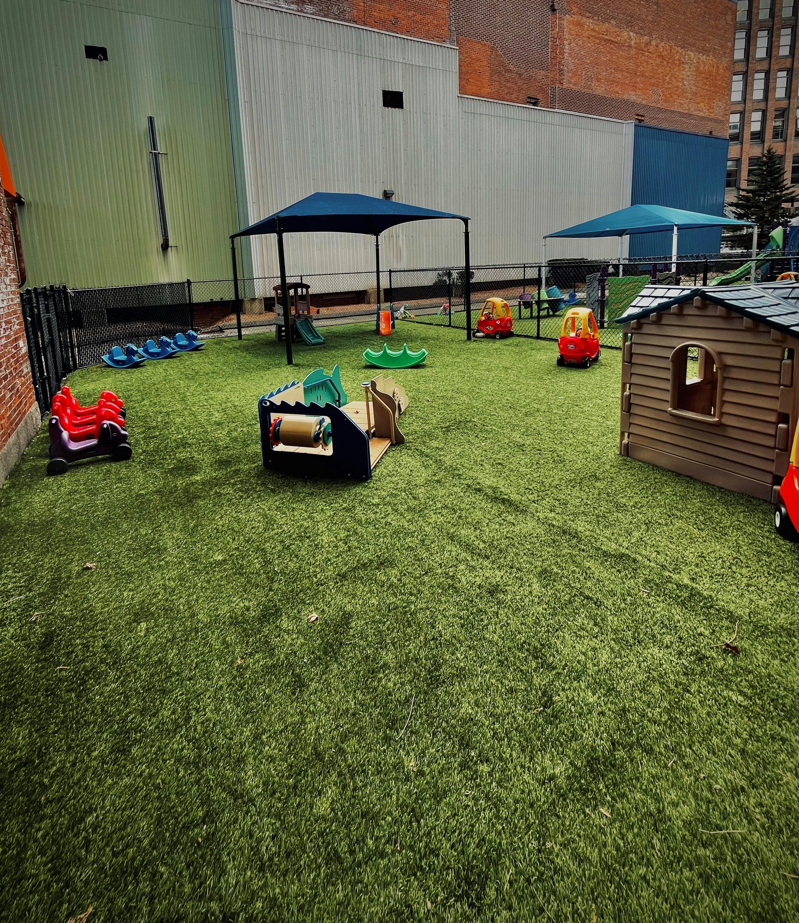 A fenced-in outdoor play area with artificial turf, playhouses, and riding toys
