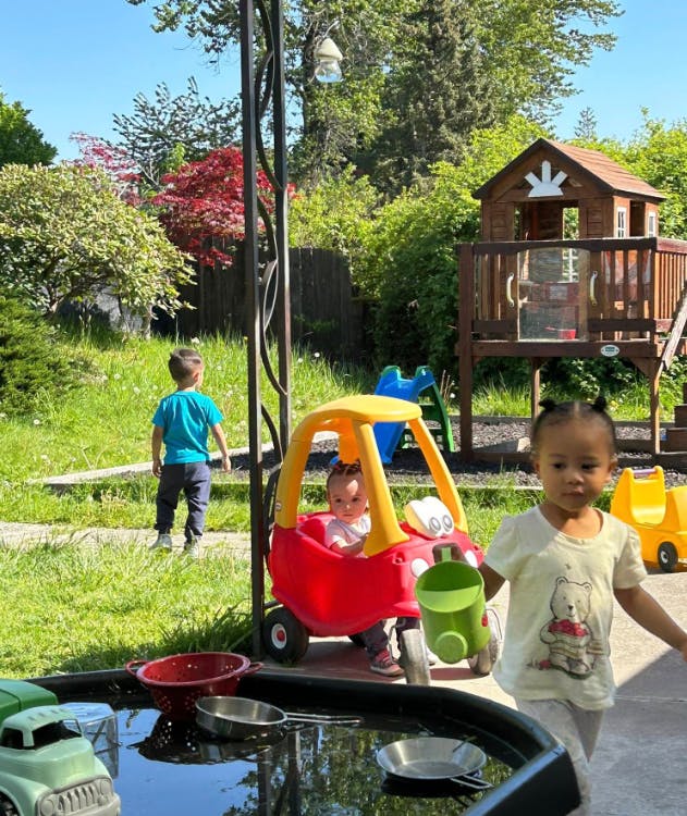 Three children playing outdoors with toys and a playhouse