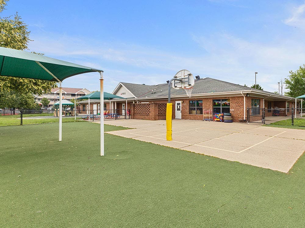 Outdoor play area with a green turf ground, shade structures, and a basketball hoop