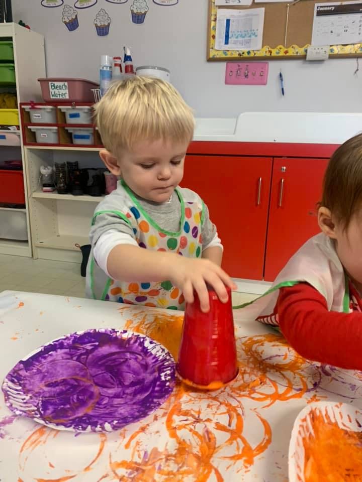 Child in an apron painting with a red cup and purple cup