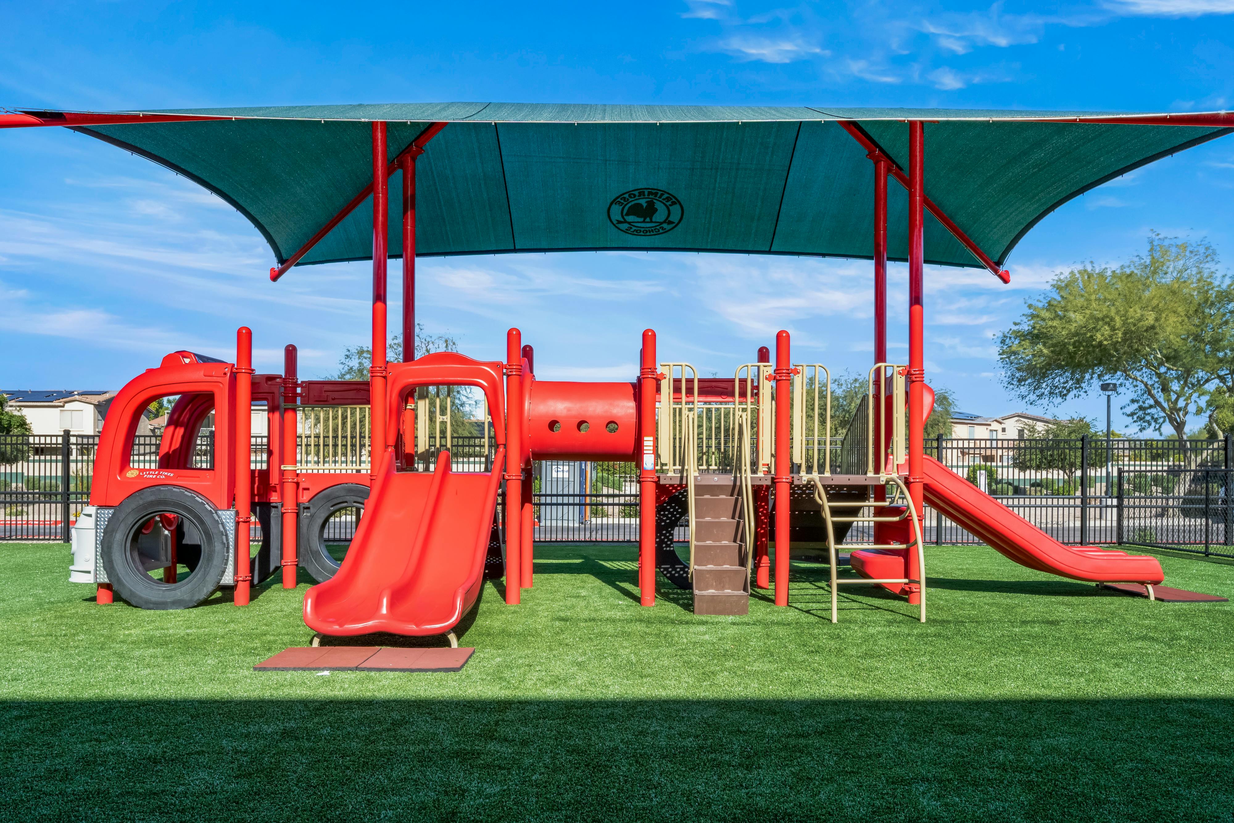 Red playground with slides, climbing structures, and a green canopy over artificial turf