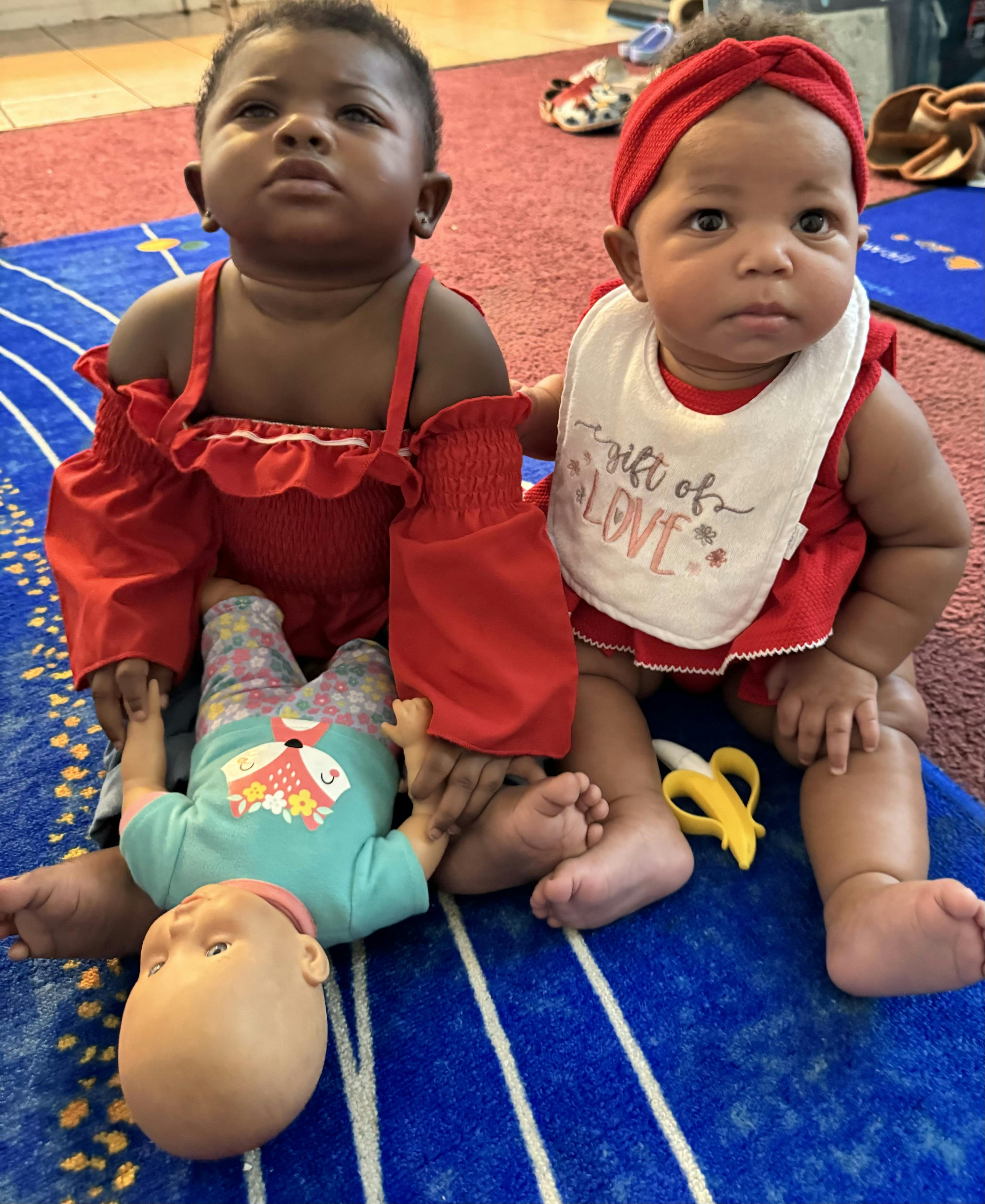 Two infants sitting on a blue rug, one holding a doll