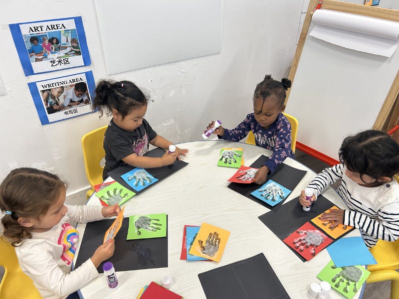 Four children at a table gluing paper handprints onto black paper in a classroom