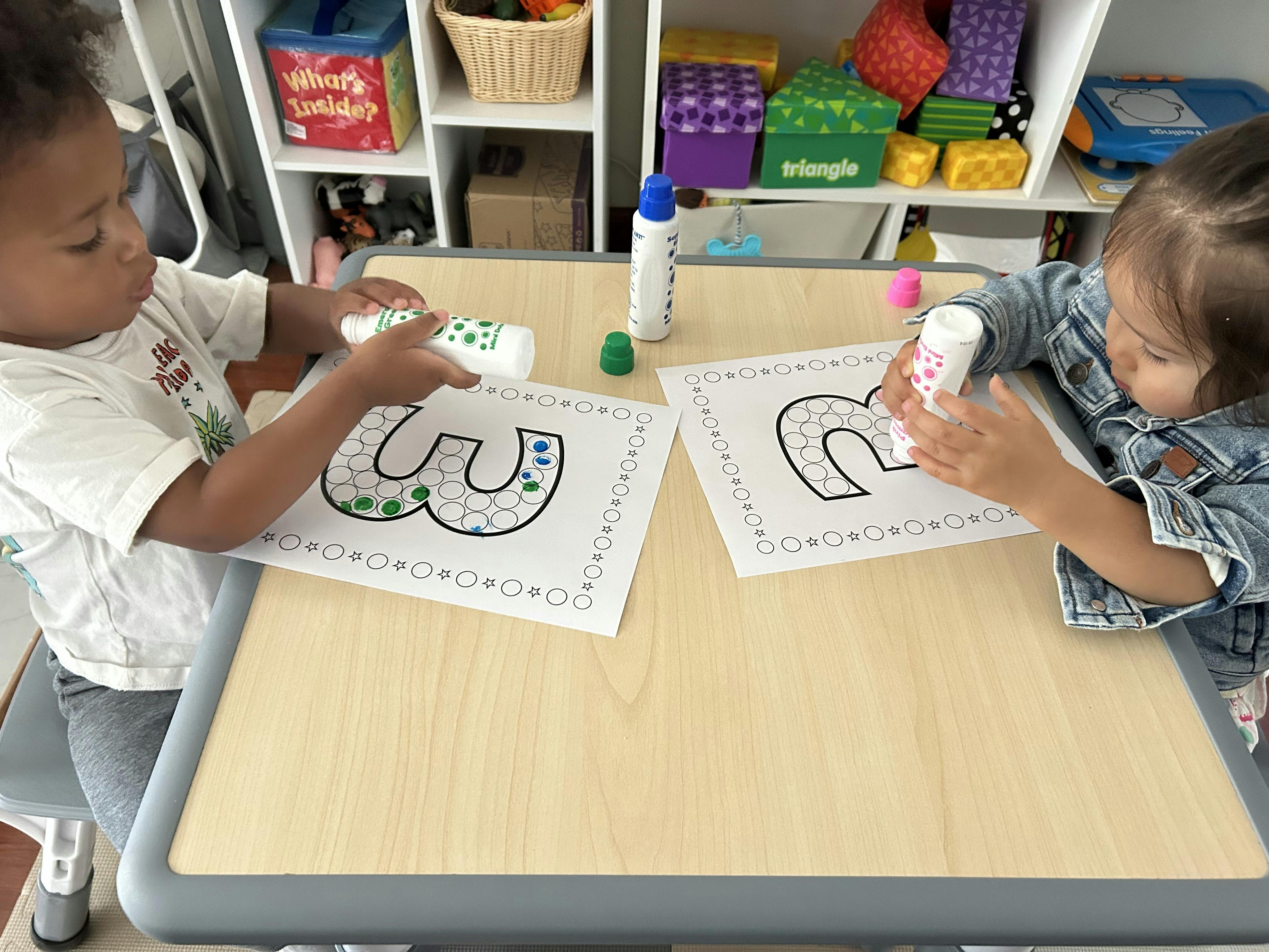 Two children at a table using dot markers to color in paper letters