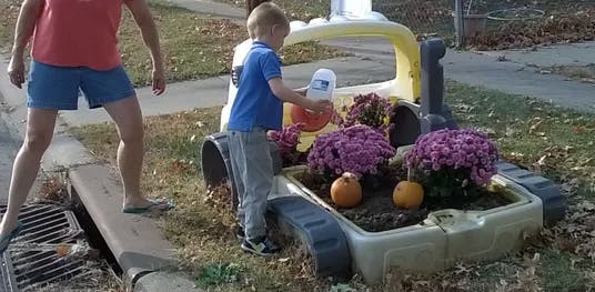 Person standing on curb next to child watering flowers in toy truck