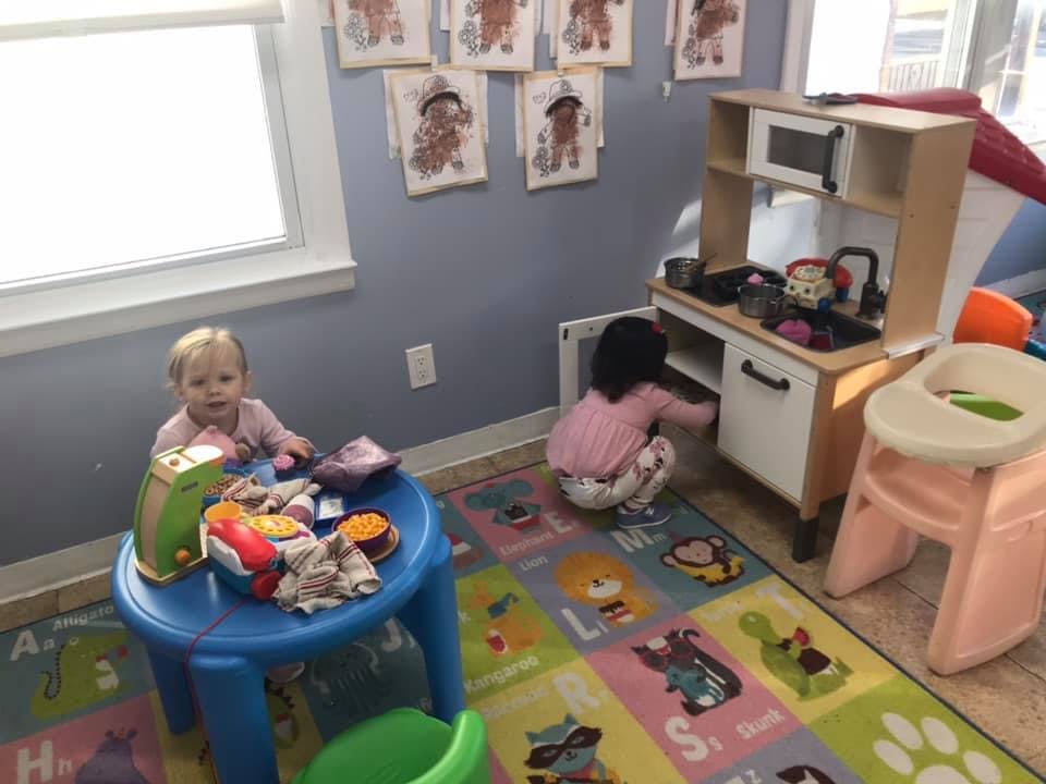Two children playing in a room, one at a table and one at a toy kitchen