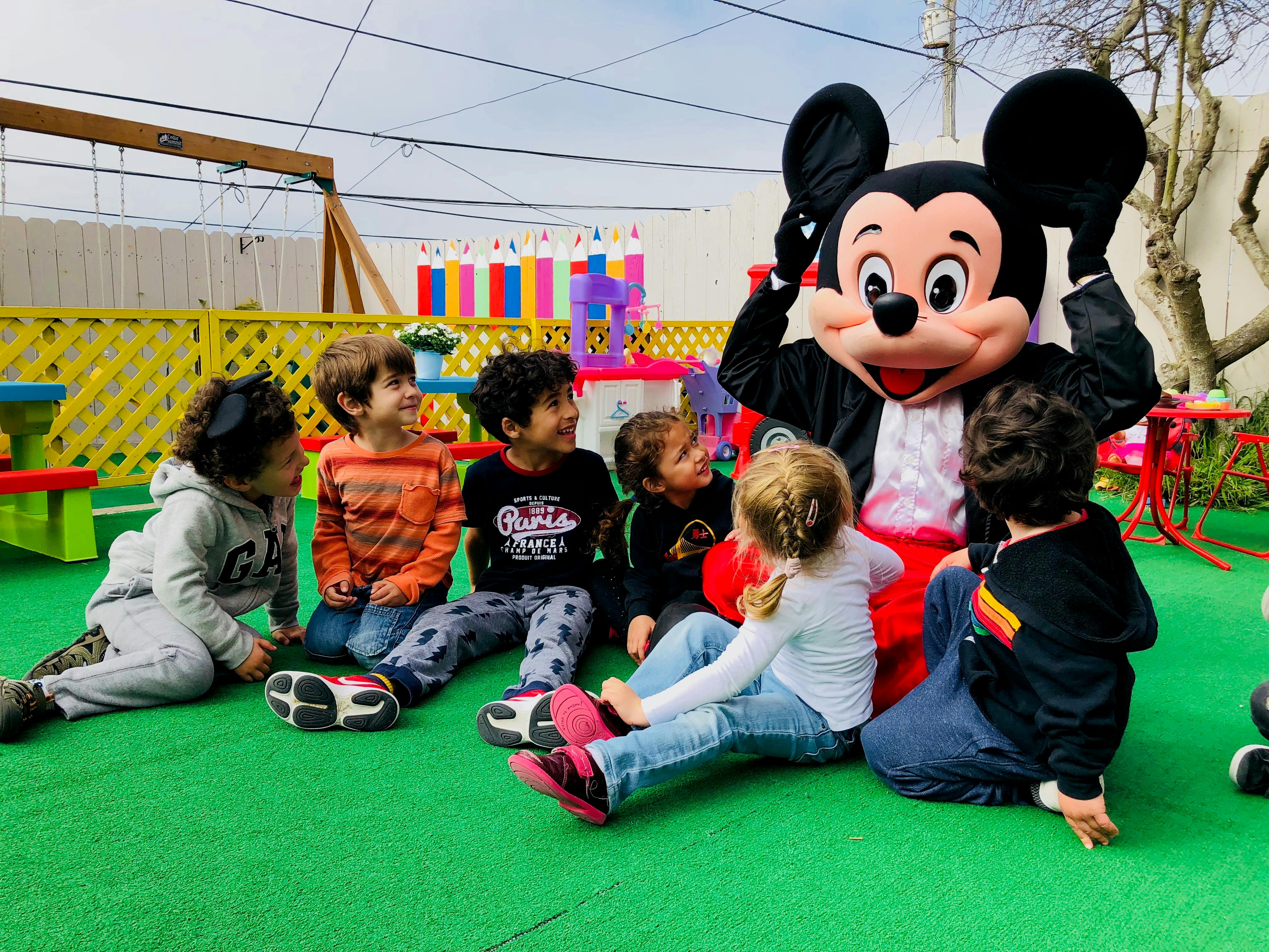 Children sitting on the ground with a person in a Mickey Mouse costume