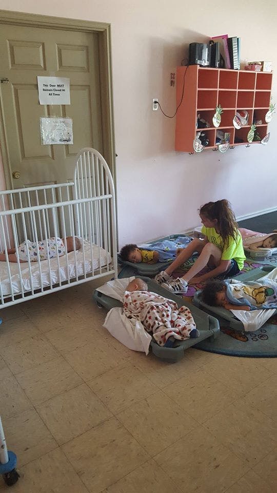 Child sitting on floor next to sleeping children on cots and a baby in a crib
