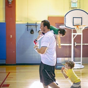 Adult carrying a child on shoulders in a gym, another child nearby