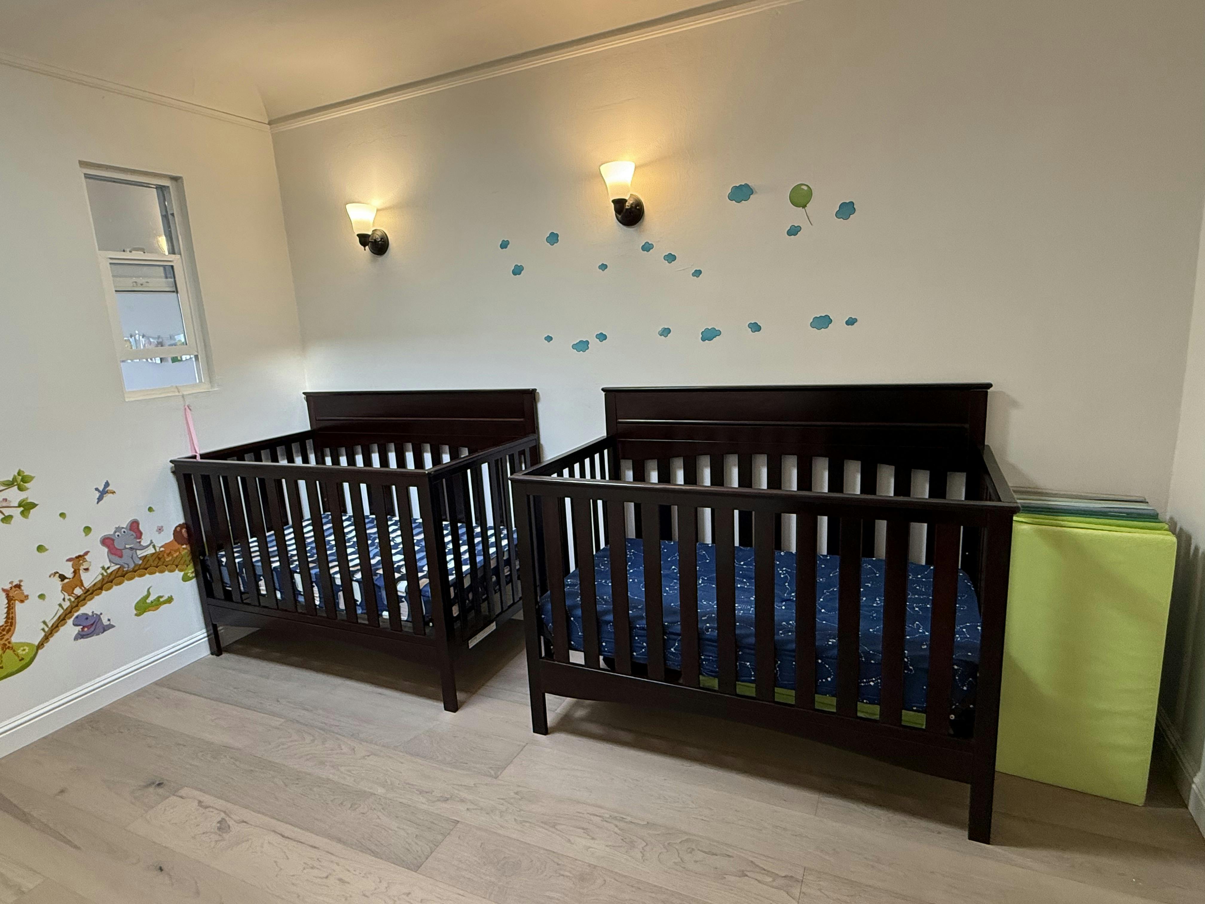 Two dark wood cribs with blue bedding in a room with wall decals