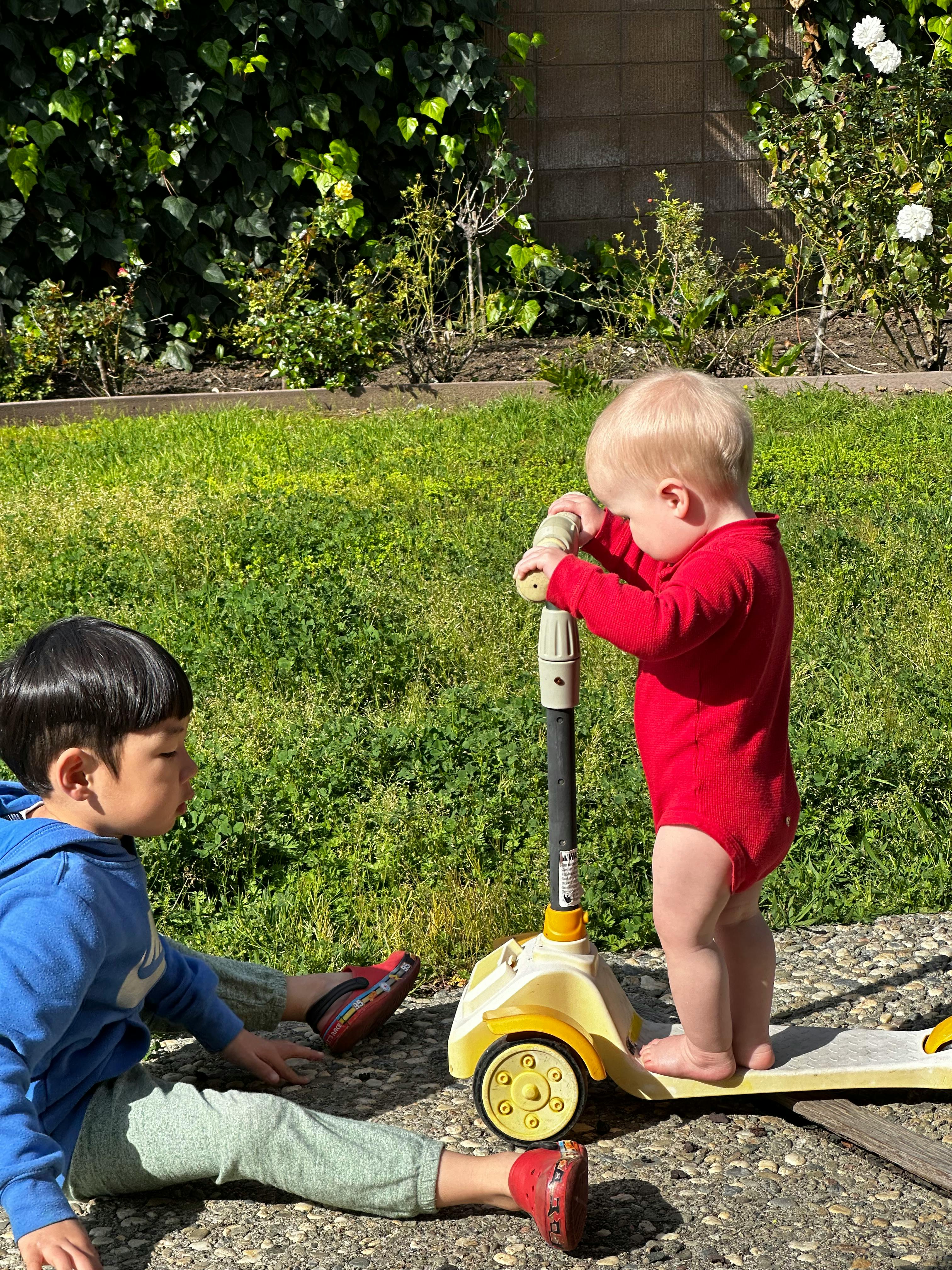 Child sitting on ground next to a baby standing on a scooter