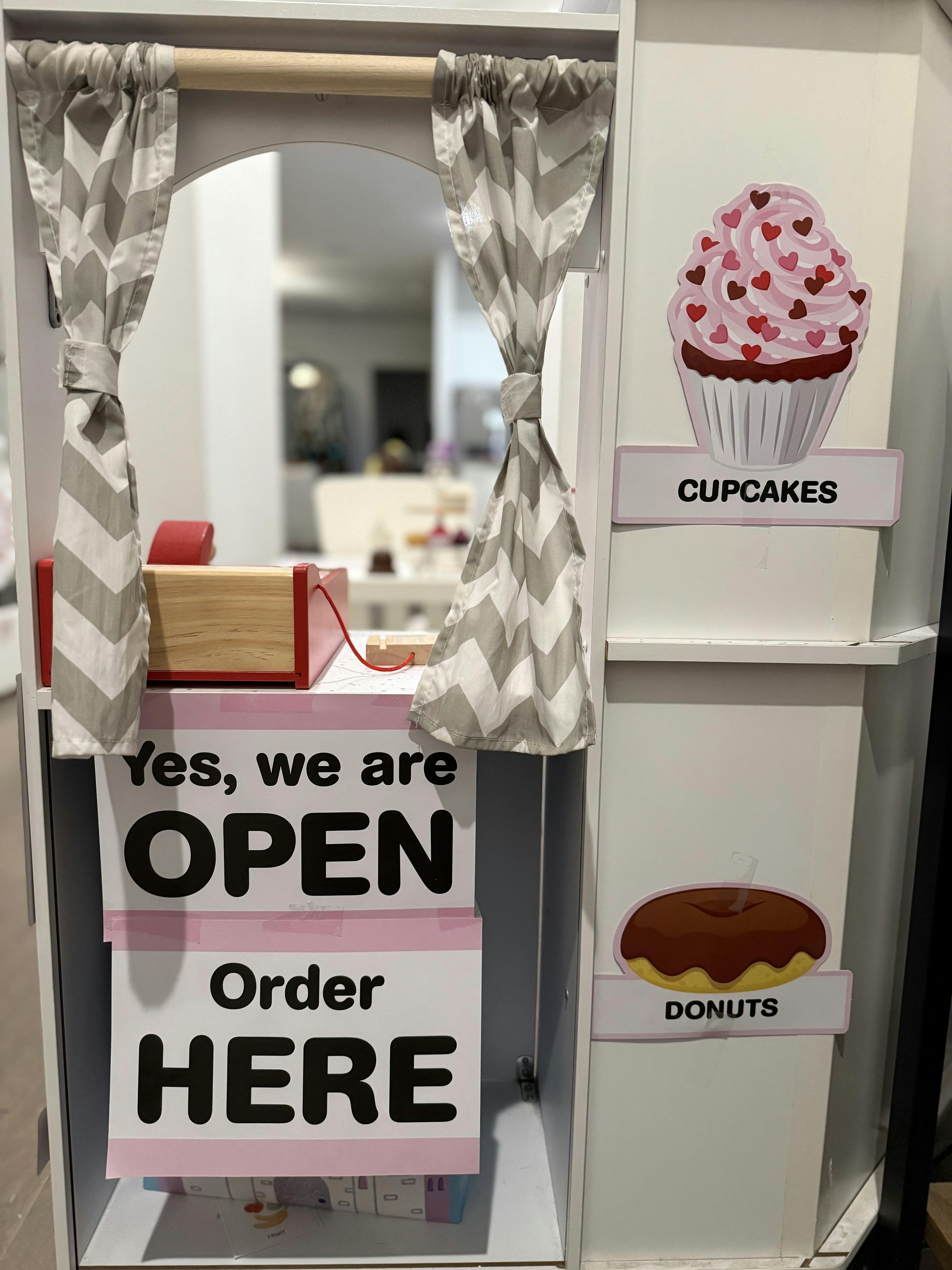 Play store with "Yes, we are OPEN Order HERE" sign, cupcake, and donut