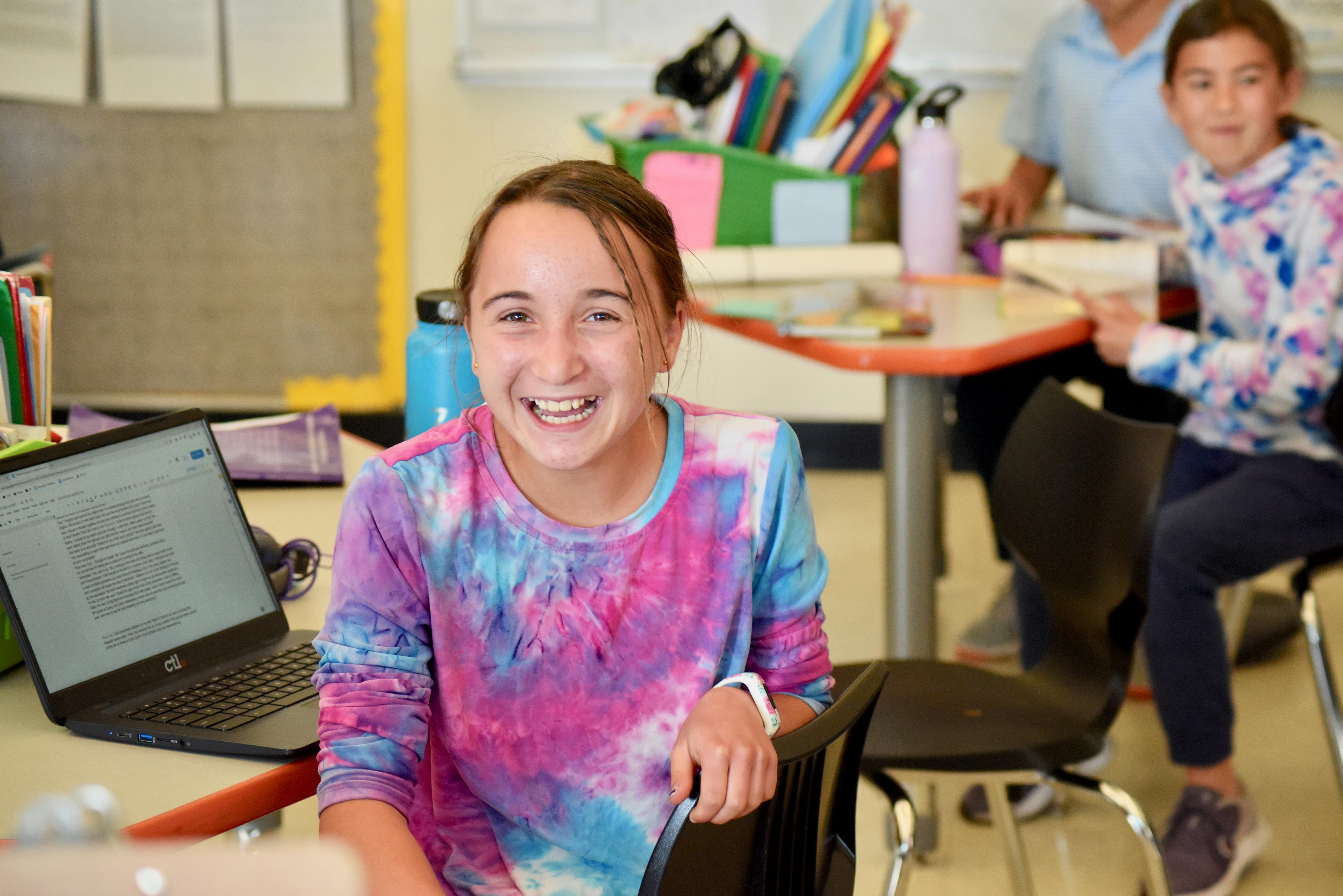 Child in tie-dye shirt smiling in a classroom with a laptop nearby
