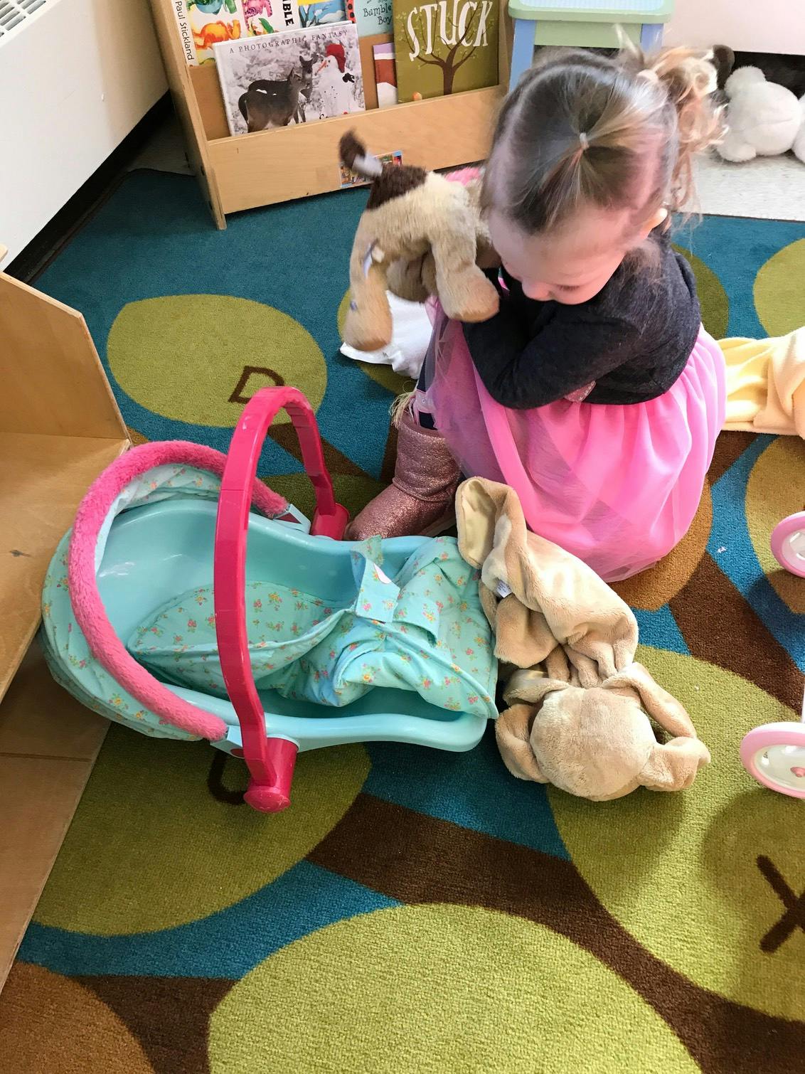 Child playing with a stuffed animal and a doll carrier on a patterned rug