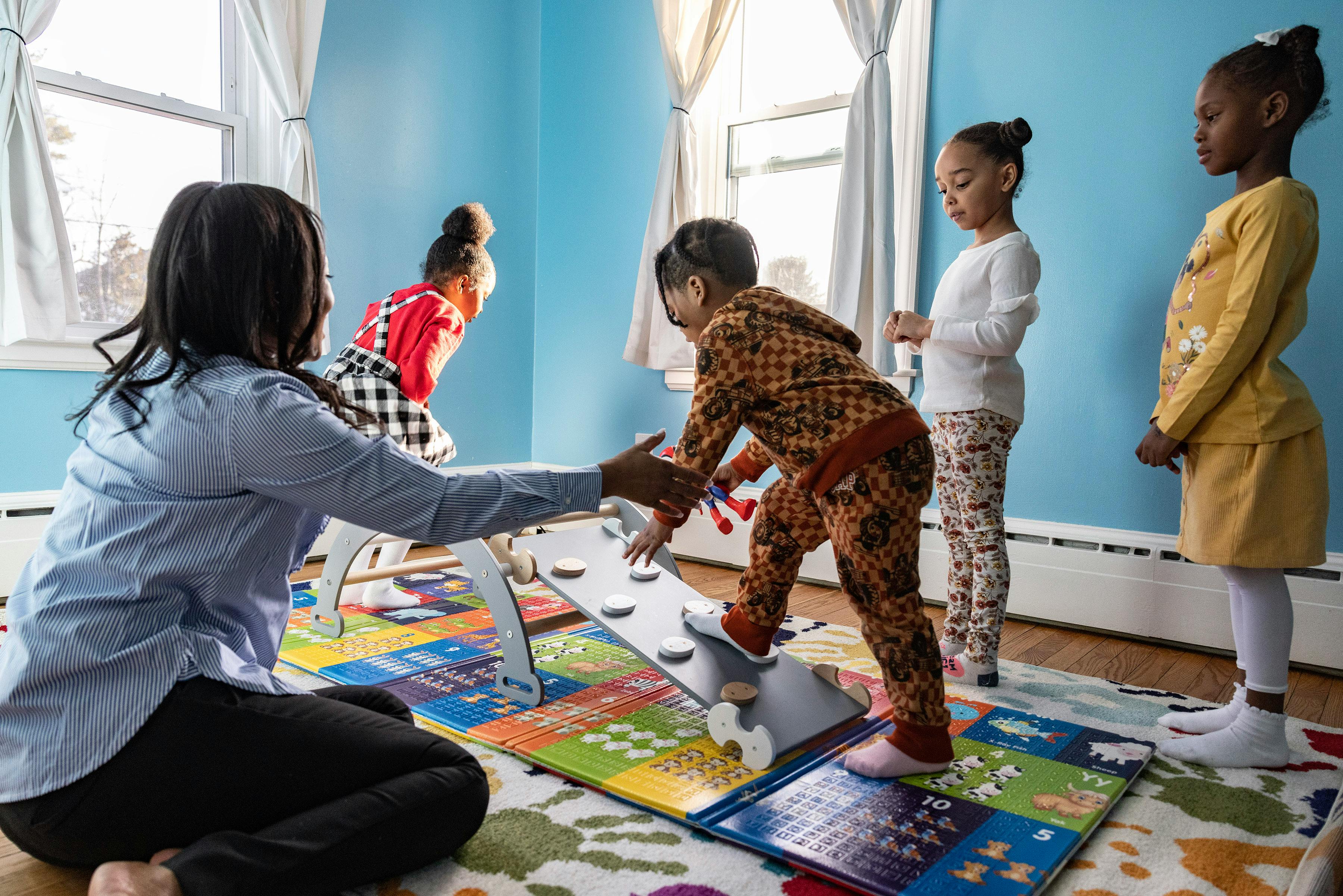Person helping a child walk on a balance beam while other children watch