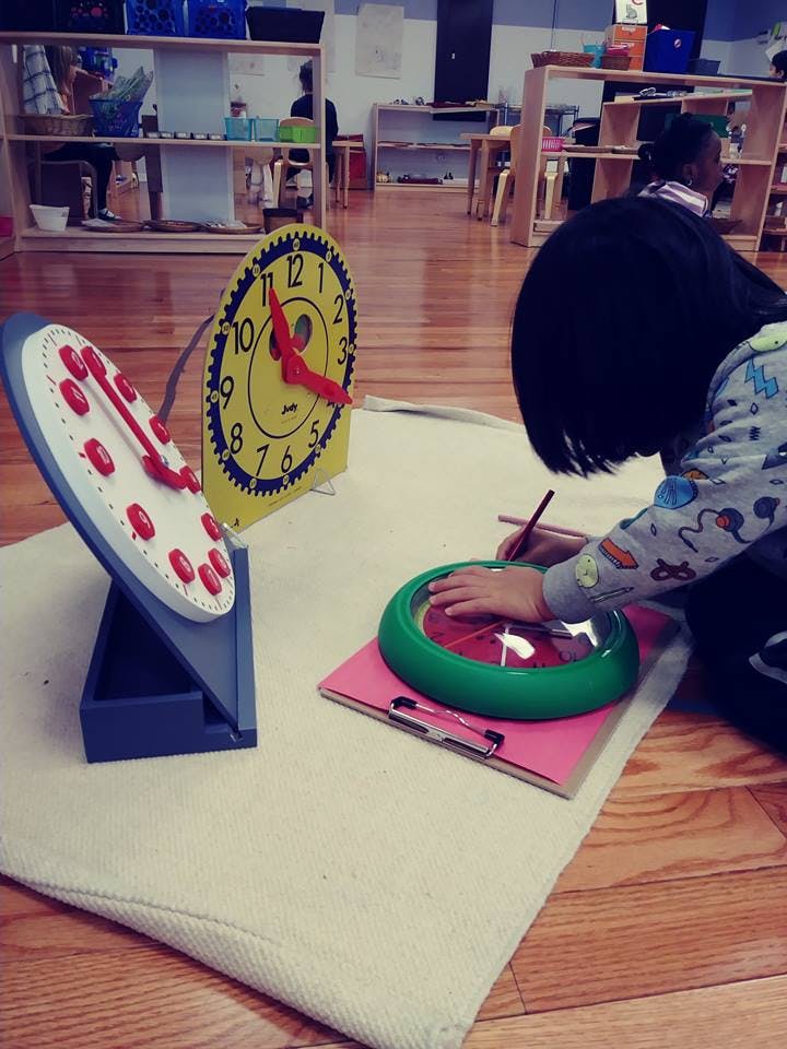 Child drawing on paper with a clock toy in a classroom