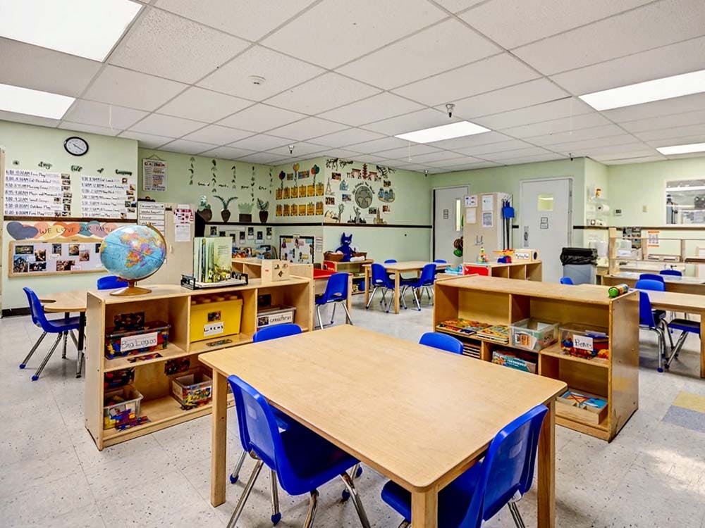 Classroom with small wooden tables, blue chairs, and toy shelves