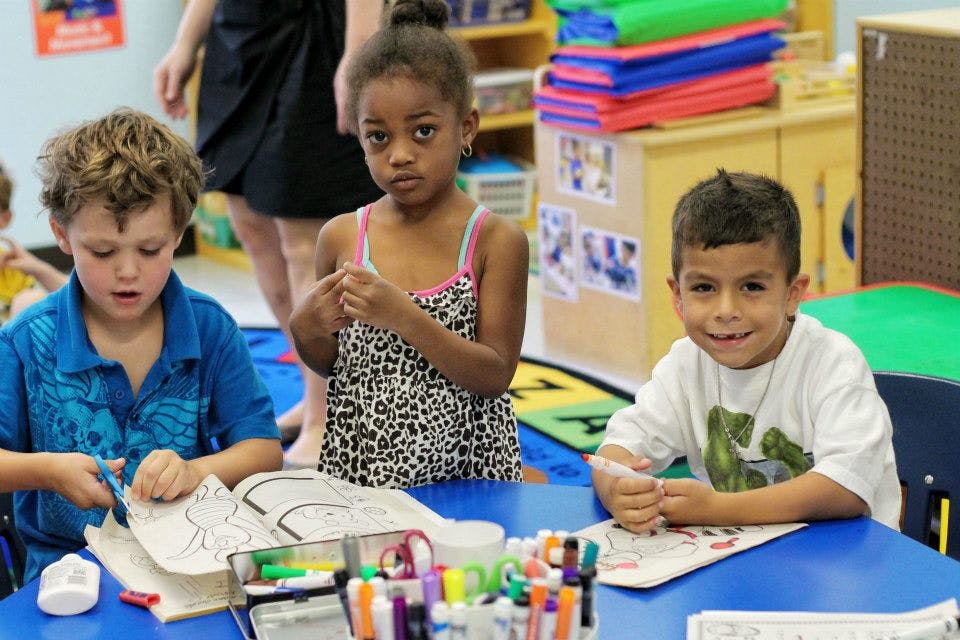 Three children at a table, one cutting paper, one looking at the camera