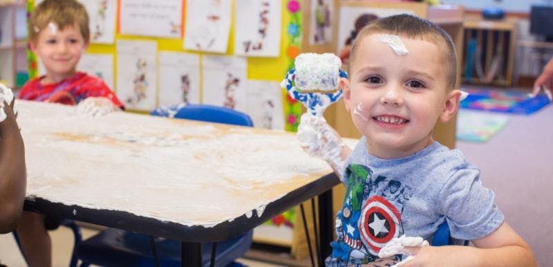 Child with foam on face and hands holding a foam-covered roller in a classroom