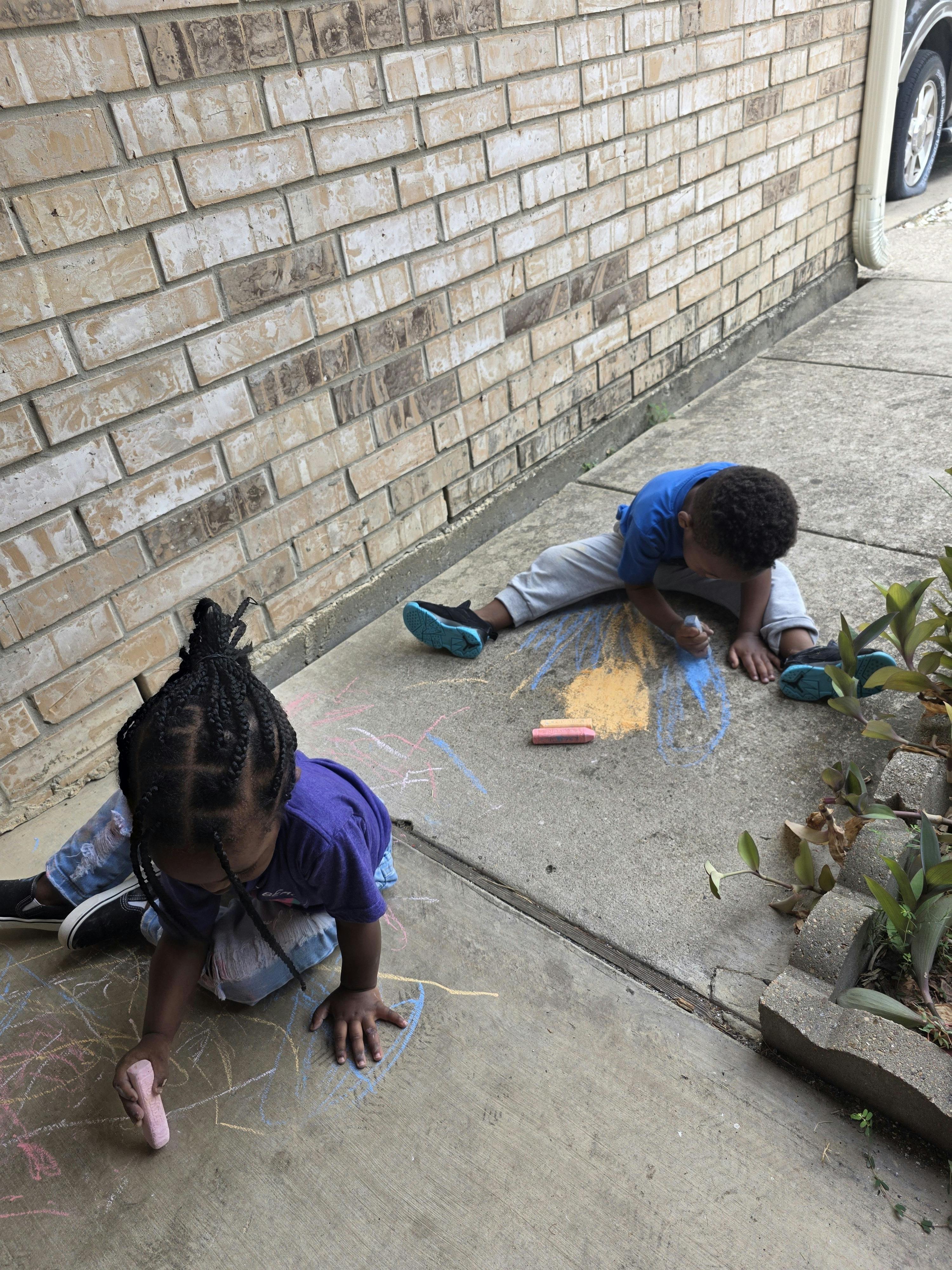 Two children drawing with chalk on a concrete sidewalk next to a brick wall