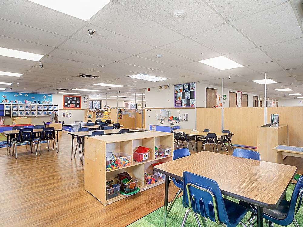 Classroom with small tables and chairs, wood floors, and shelves with toys