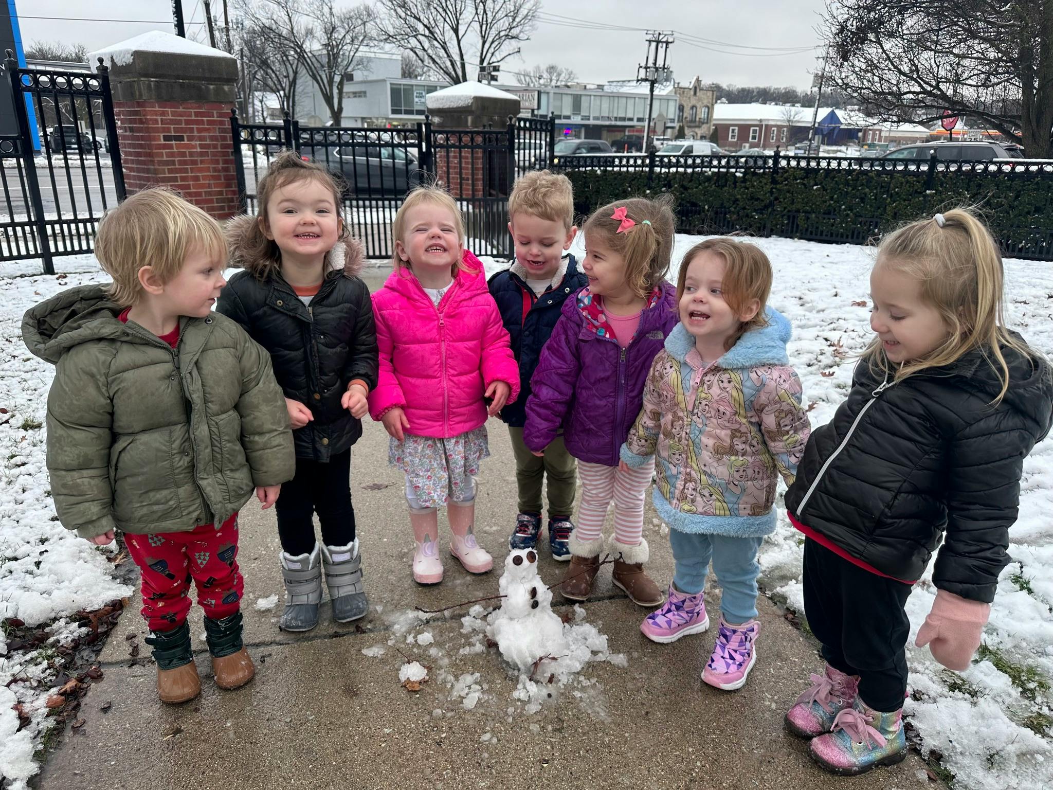 Six children standing around a small snowman in the snow