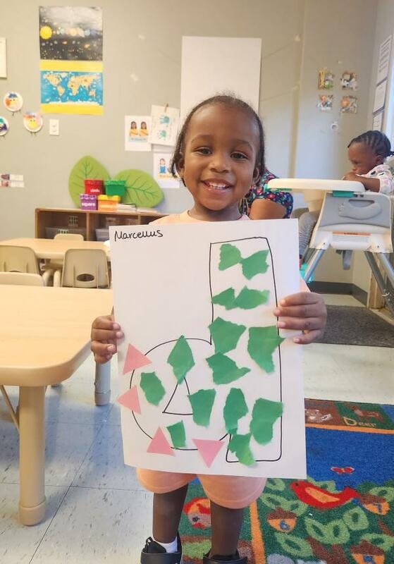 Child holding up a paper with a letter J and number 4 decorated with shapes
