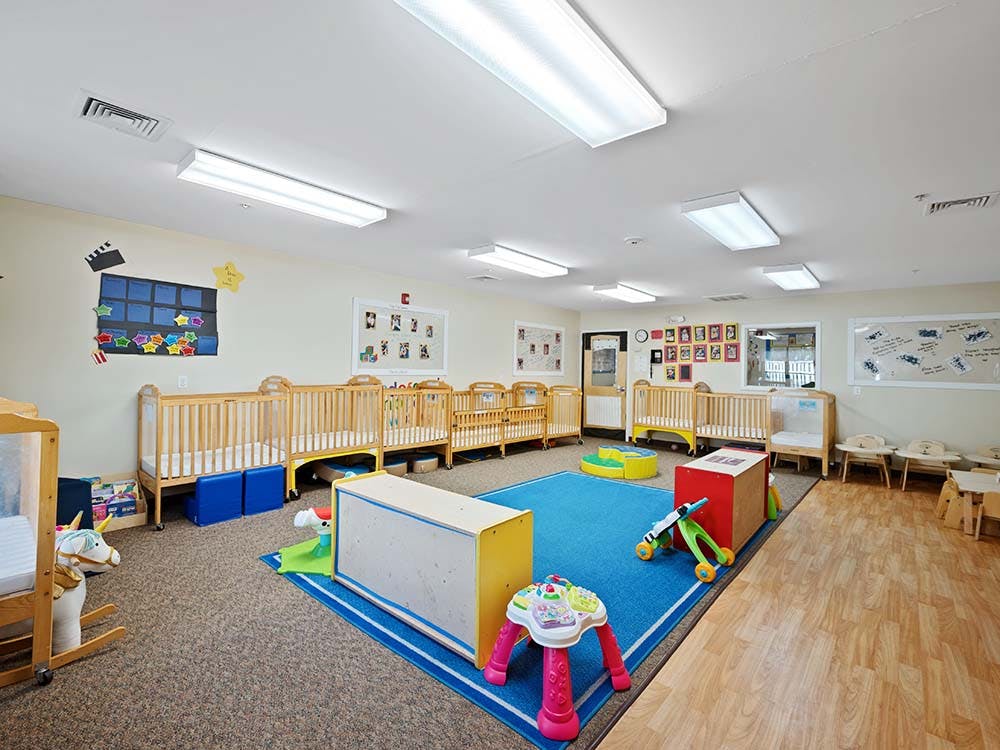 A brightly lit nursery room with multiple wooden cribs and a blue play mat