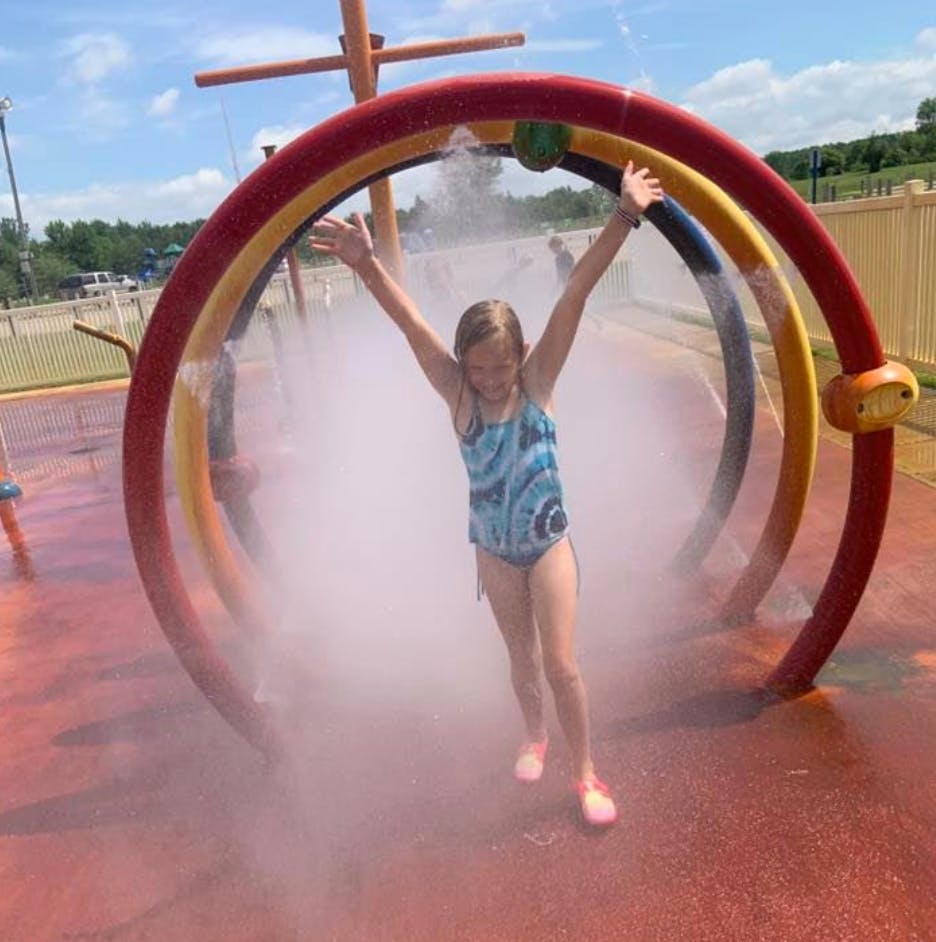 Child in a swimsuit with arms raised, standing in mist from a water play structure
