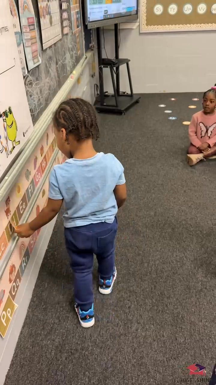 Child touching an alphabet chart on a classroom wall, another child sitting on the floor