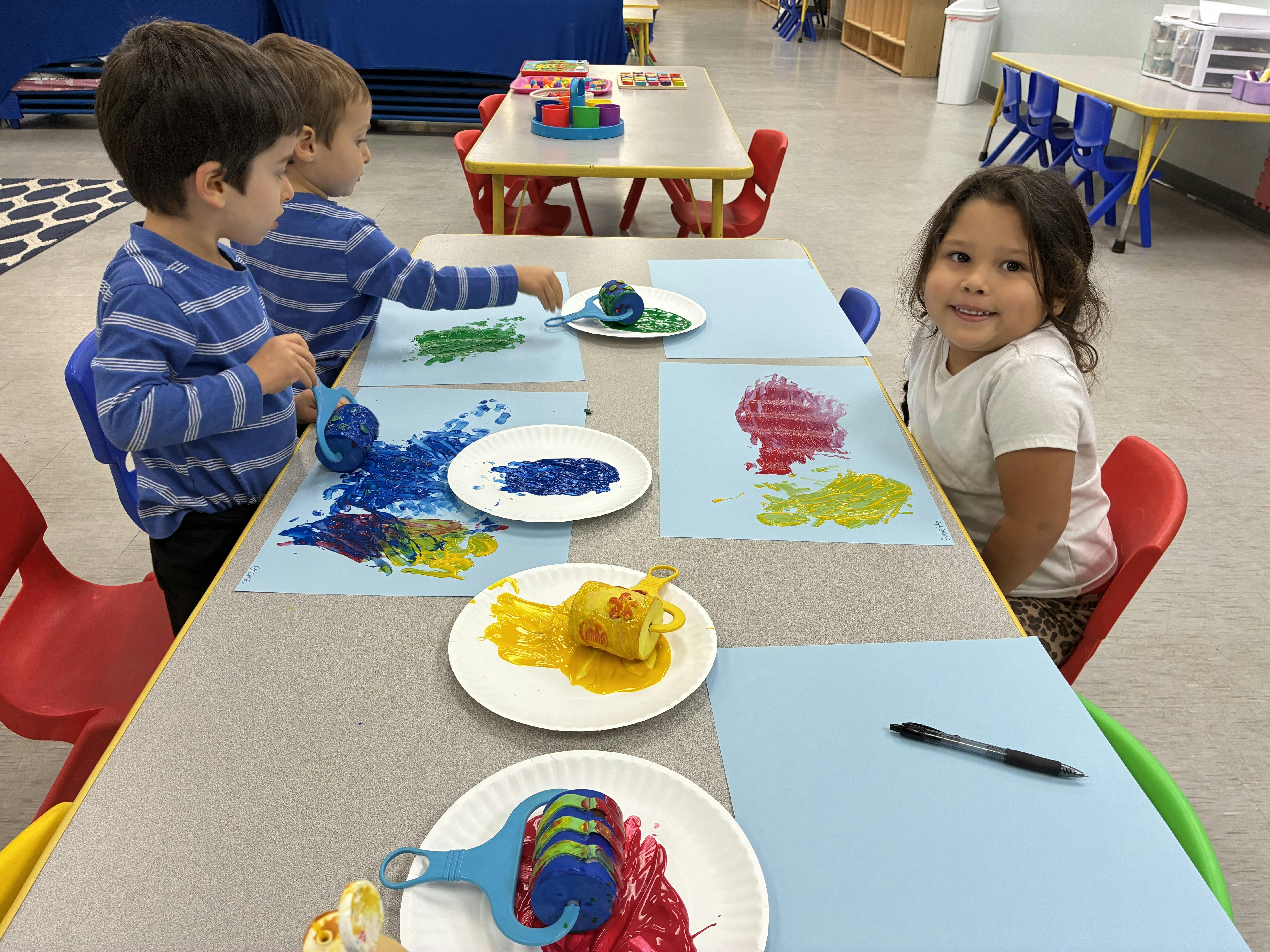 Three children painting on paper with rollers at a classroom table