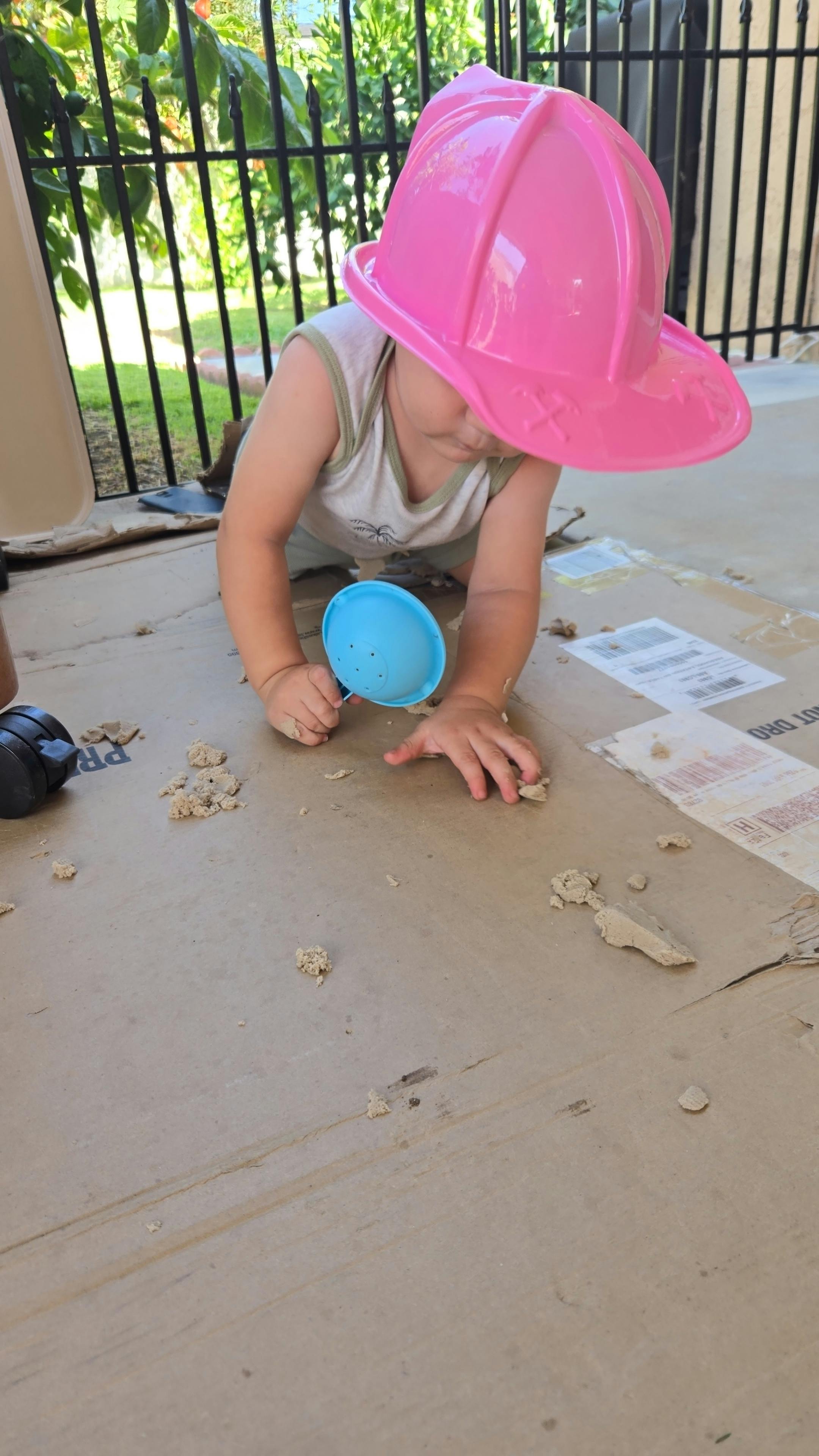 Child wearing a toy helmet playing with sand on a cardboard surface