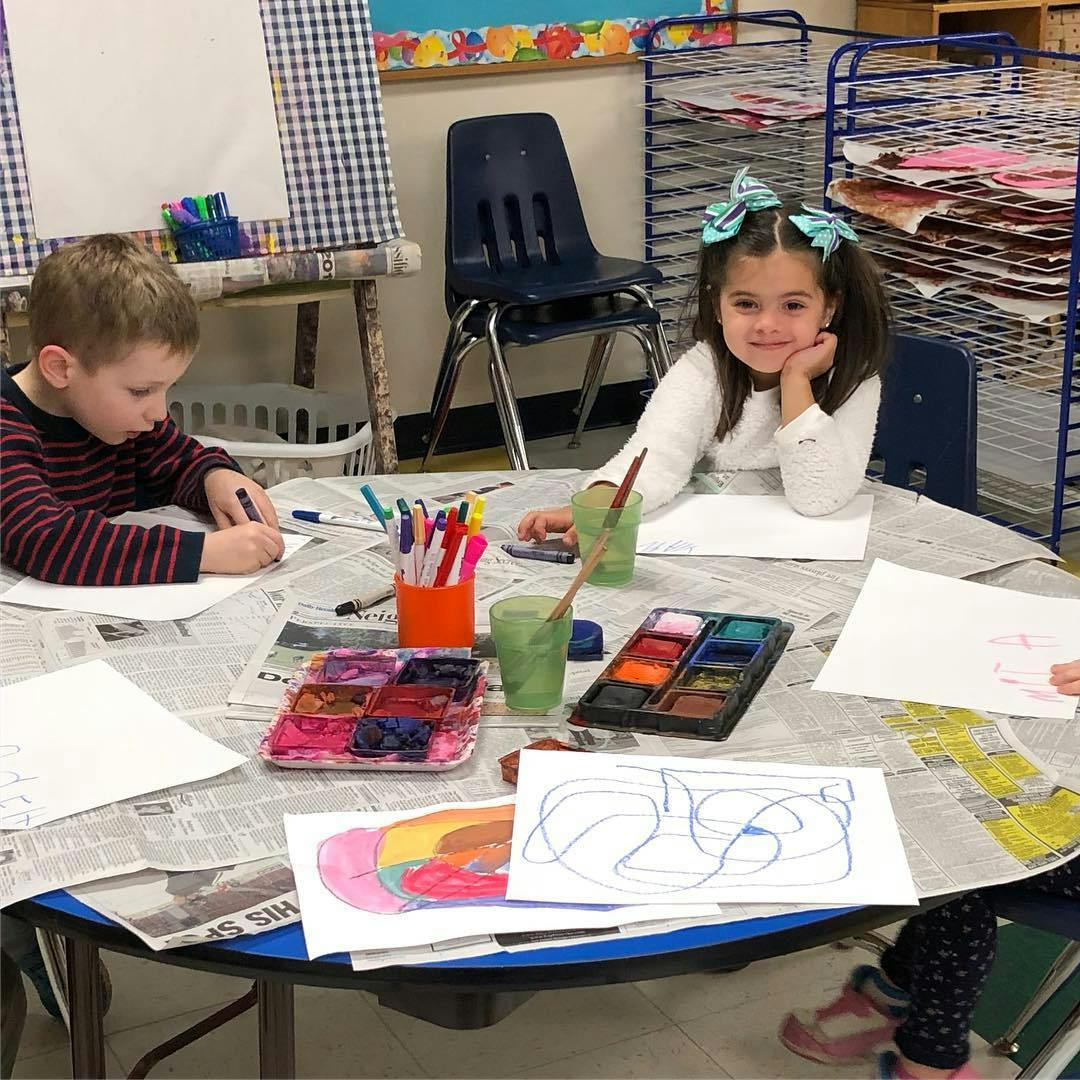 Two children painting and drawing at a table in a classroom