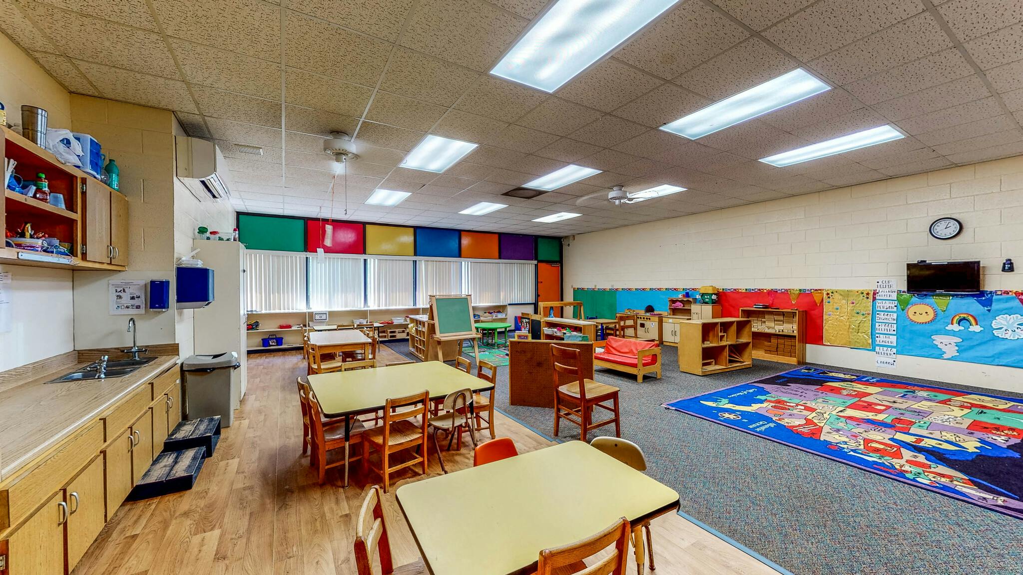 Empty classroom with small tables and chairs, a sink counter, and colorful decor