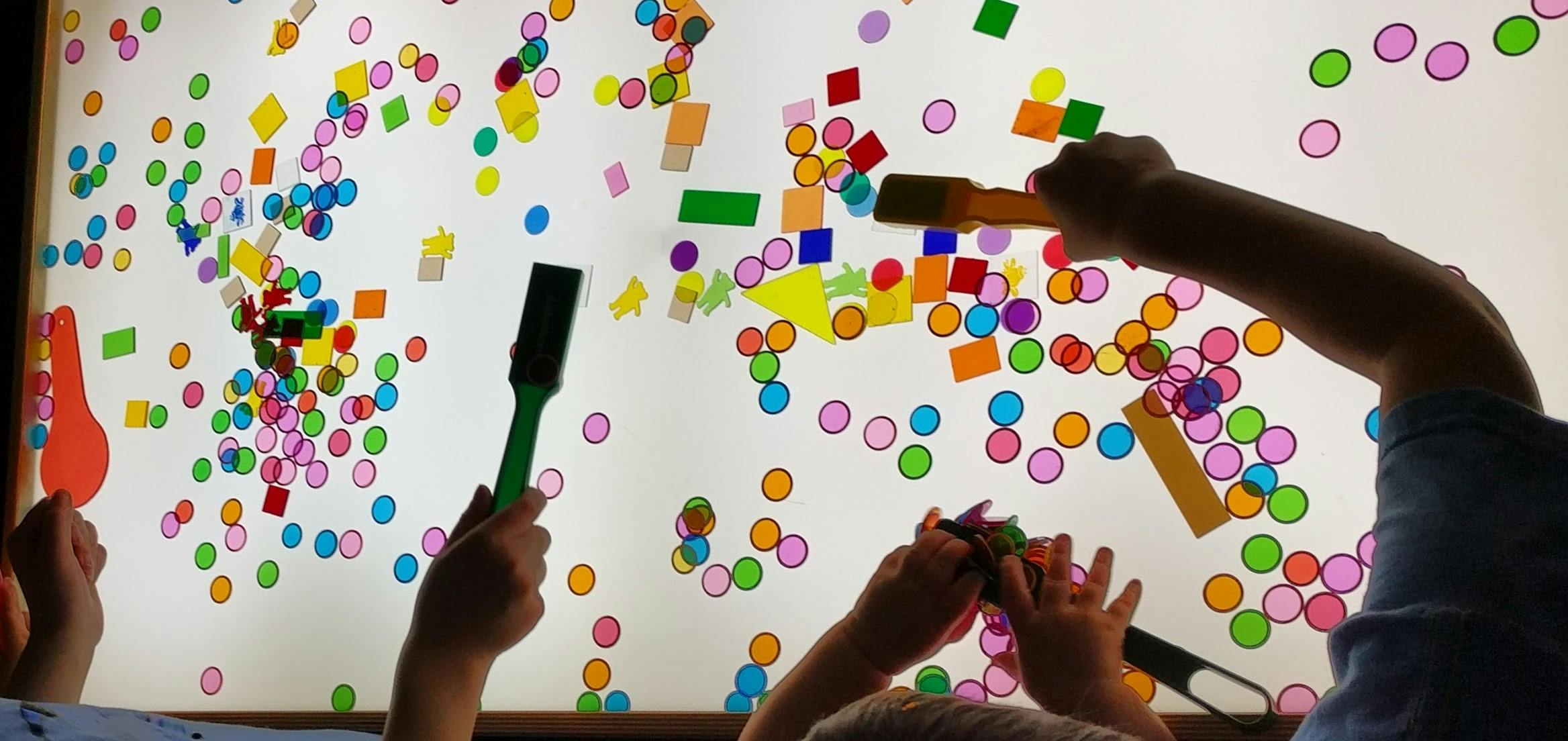 People using magnetic wands to arrange colorful geometric shapes on a light table