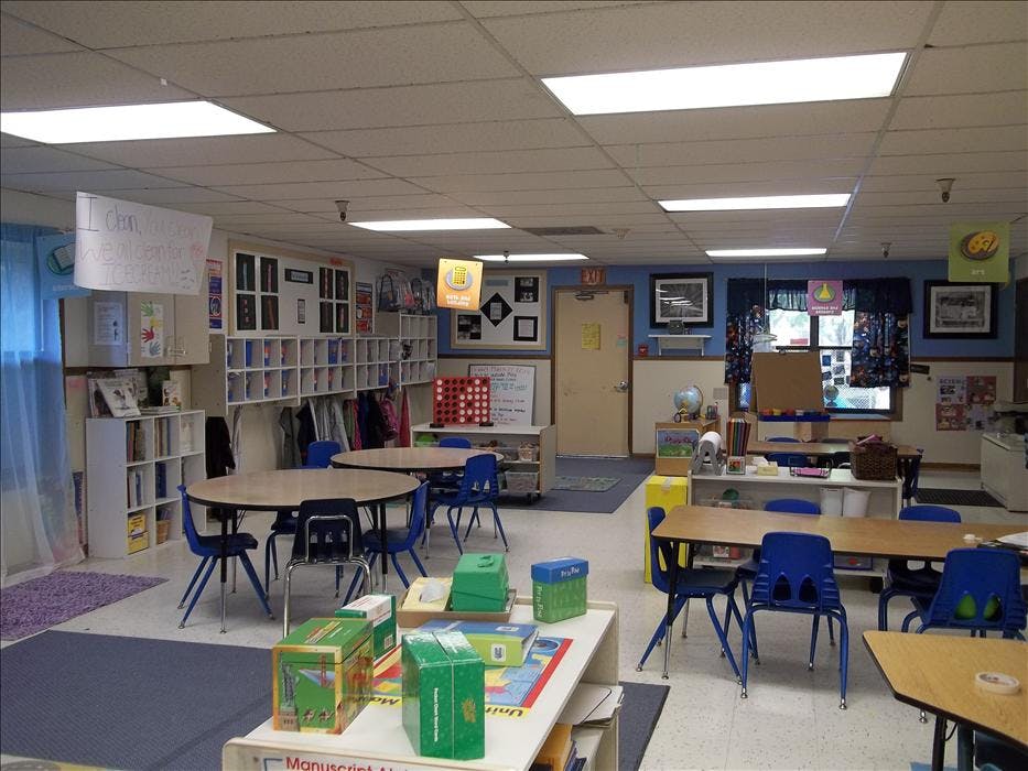 An empty classroom with several tables and chairs, shelves, and learning materials