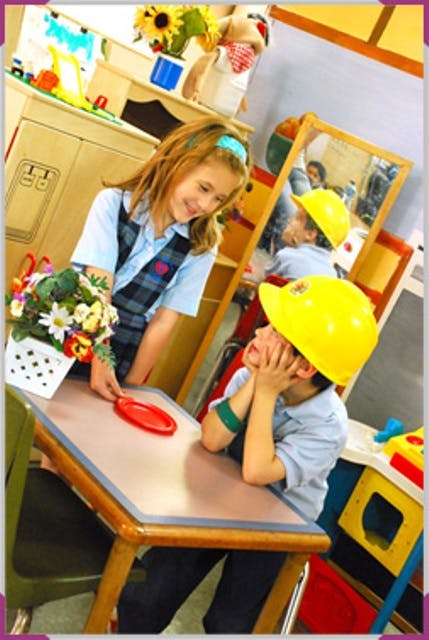 Child in uniform holding a red object over a table, child in yellow hard hat