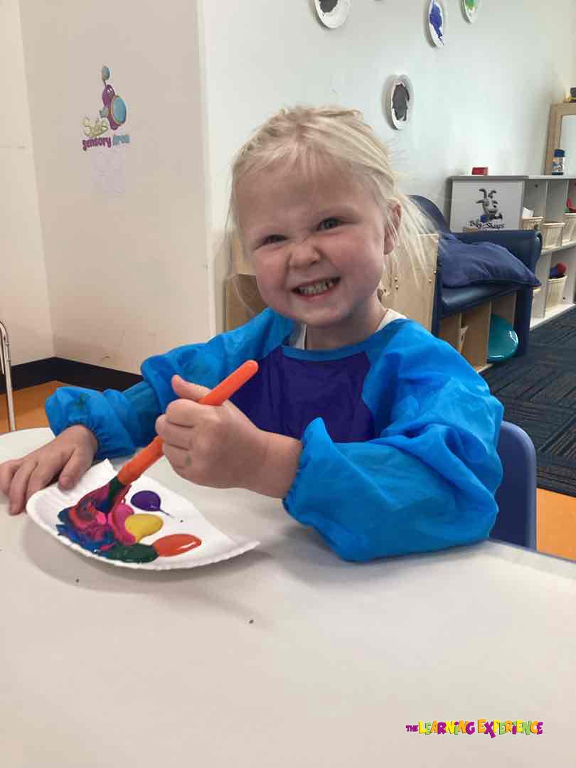 Child in a blue smock painting with a brush and colorful paints on a paper plate