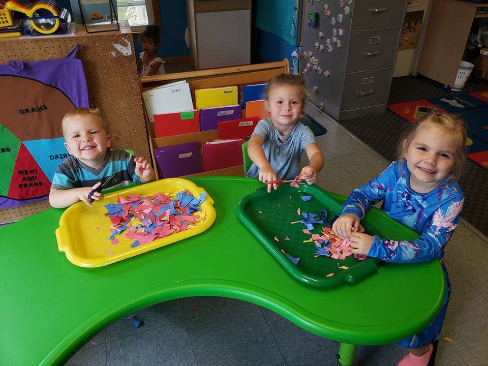 Three children at a green table with trays of colorful paper scraps