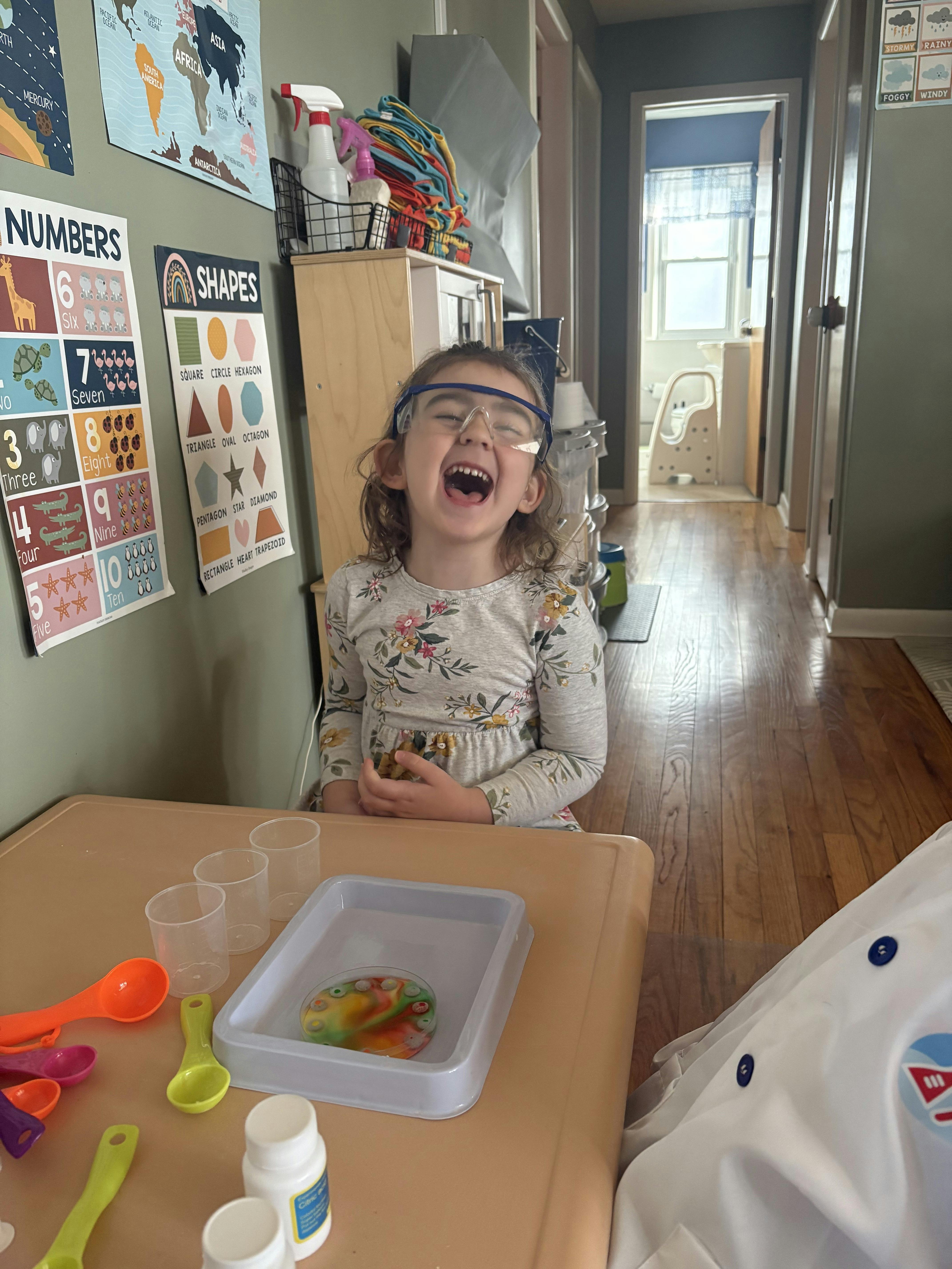 Child wearing safety goggles, laughing with mouth open, at a table with science experiment supplies