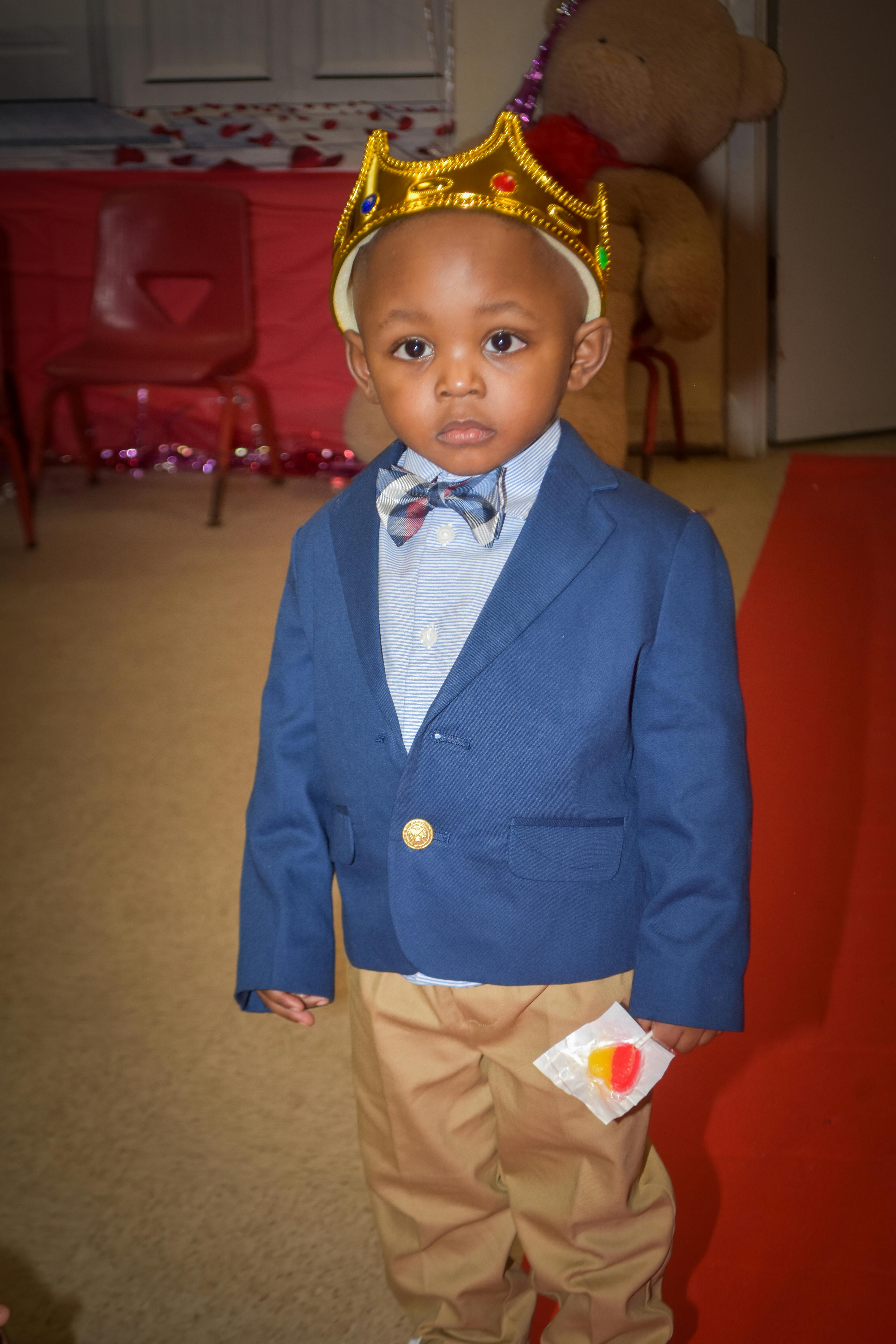 Child wearing a gold crown, blue blazer, and bow tie, holding candy