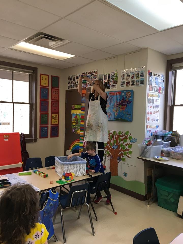 Person in apron standing on a stool, child playing with toys in a classroom
